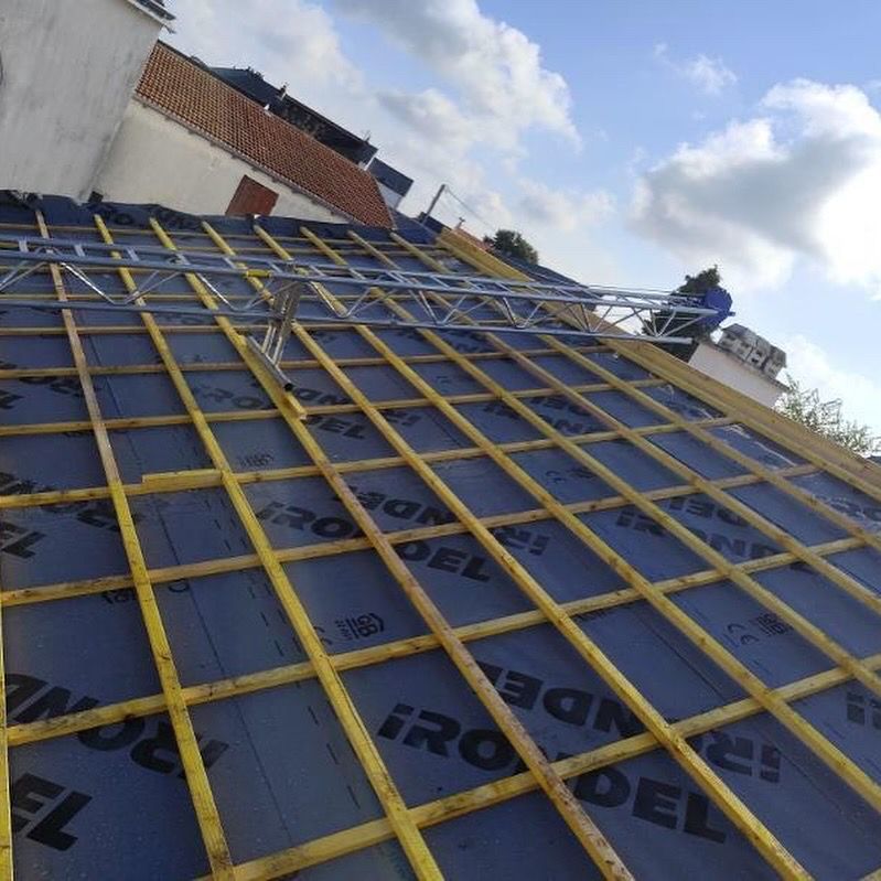 Construction du toit en cours : poutres en bois, bâche bleue, échafaudage métallique, ouvrier sur le toit sous un ciel partiellement nuageux.