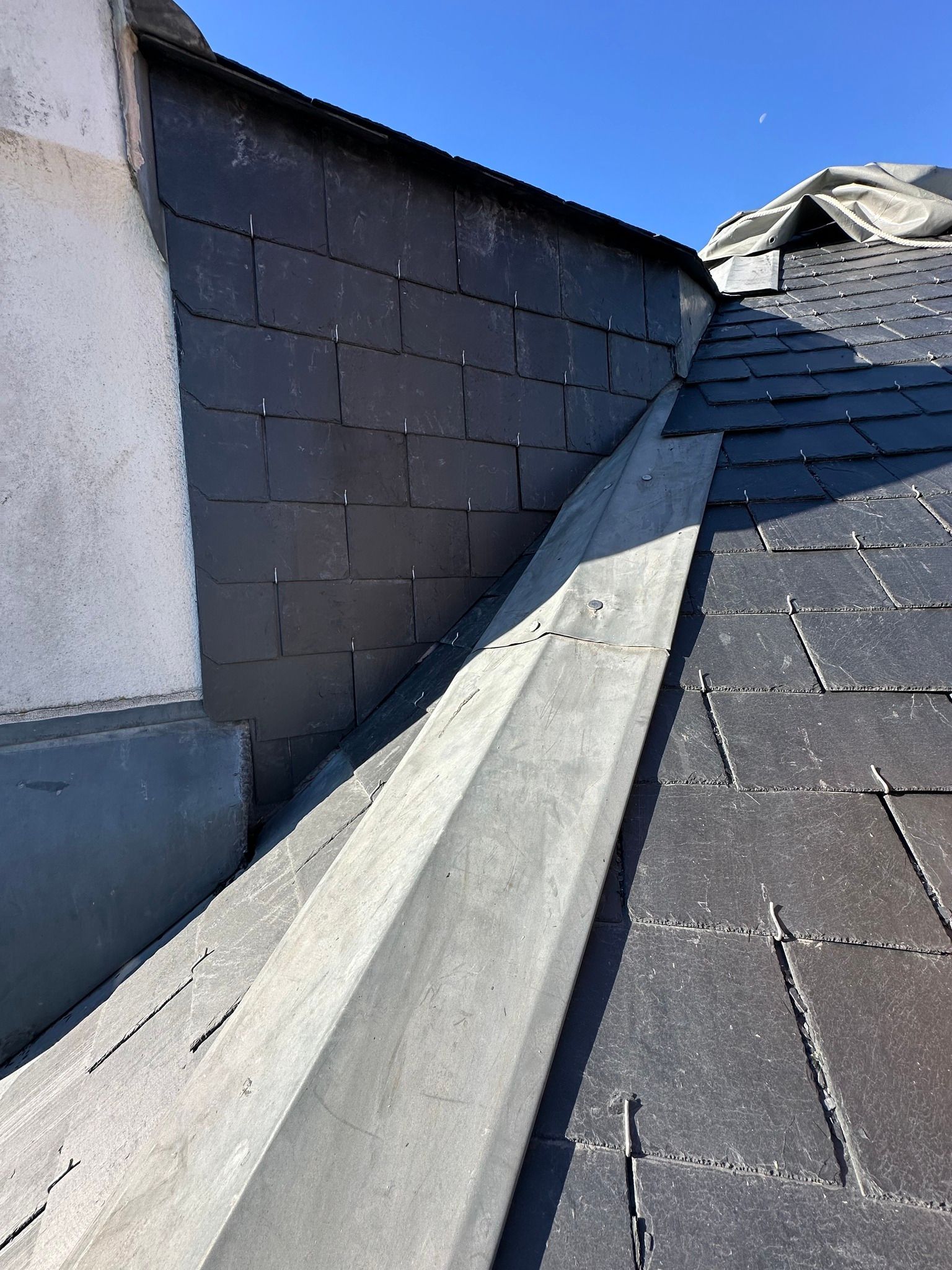 Le toit et le mur en ardoise noire se rejoignent au niveau d'une gouttière en béton gris sous un ciel bleu.