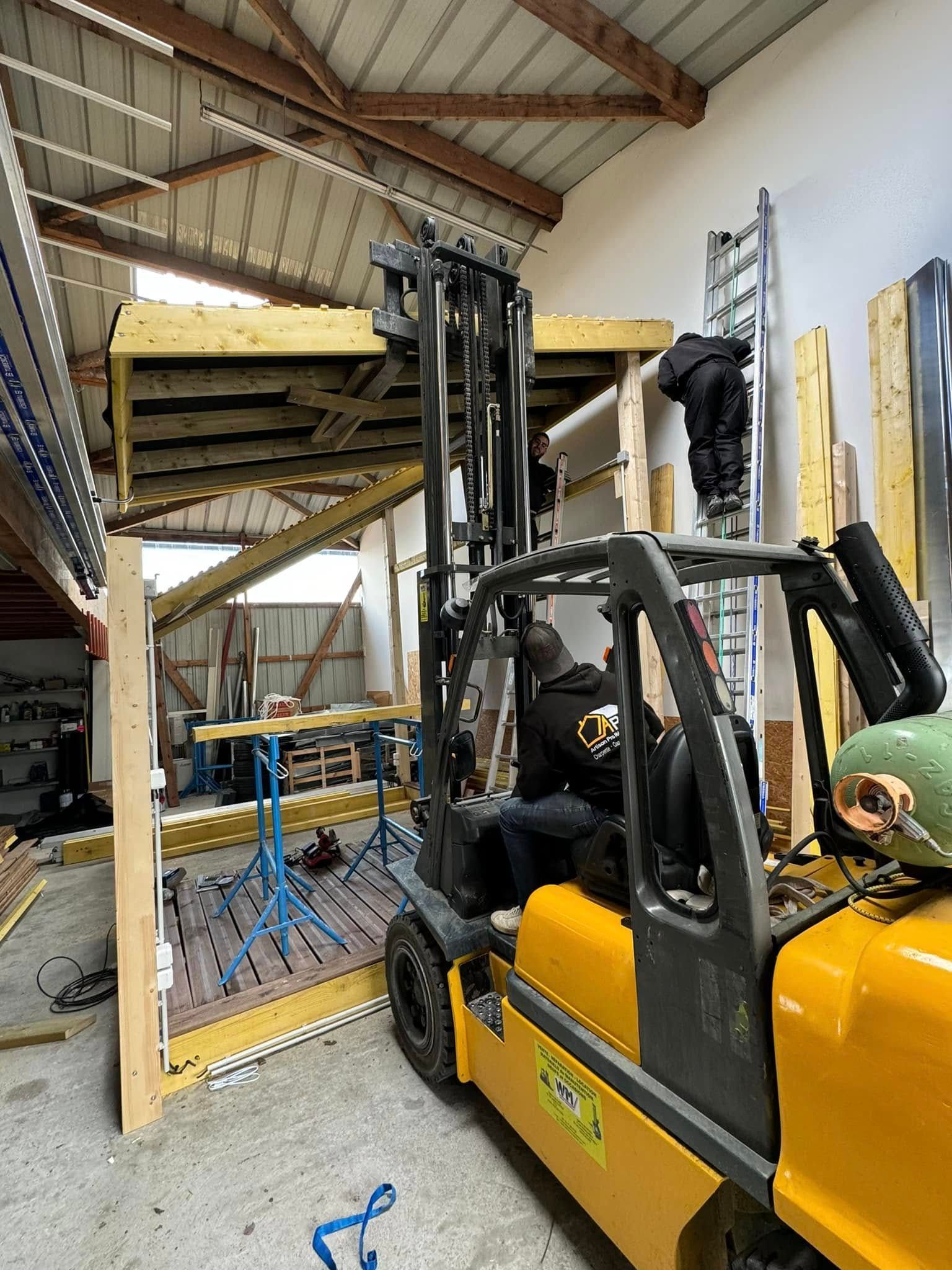 Un chariot élévateur jaune soulève du bois dans un atelier. Une personne est sur une échelle à proximité.