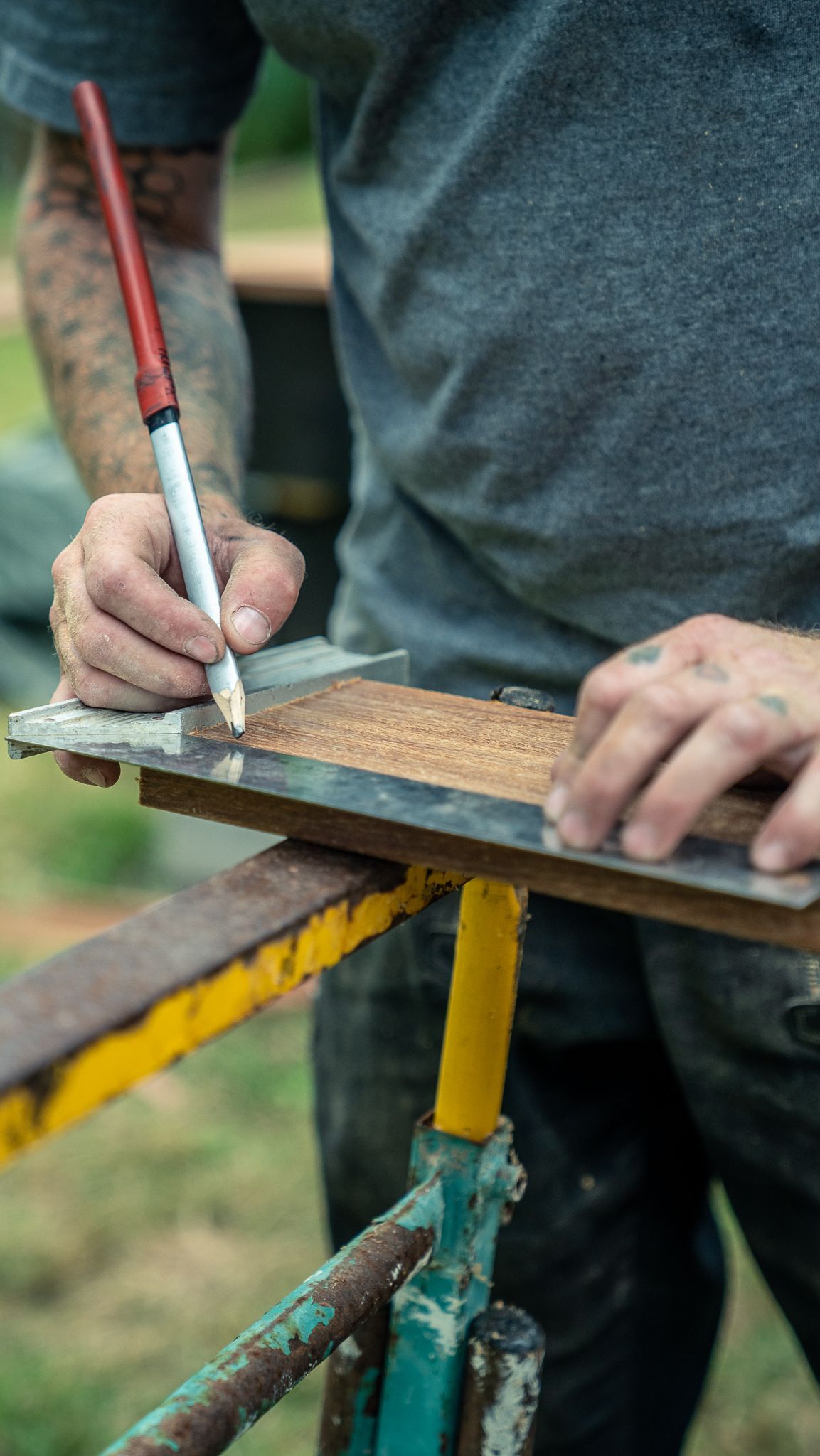 Personne utilisant un crayon et un guide en métal pour marquer le bois sur un support de travail, probablement en train de travailler le bois à l'extérieur.