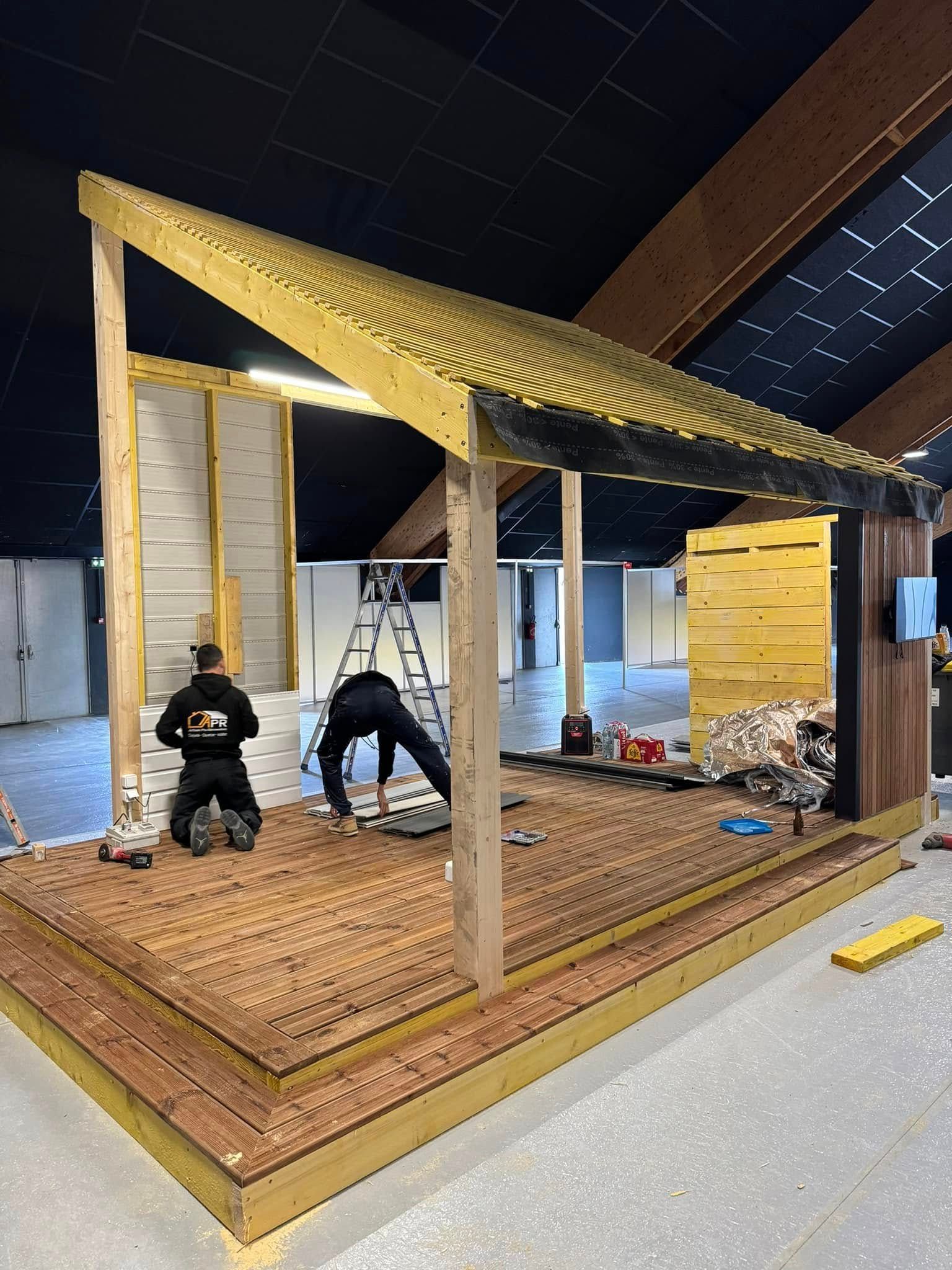 Deux ouvriers construisent une structure en bois en atelier avec une plate-forme, un toit et des murs.