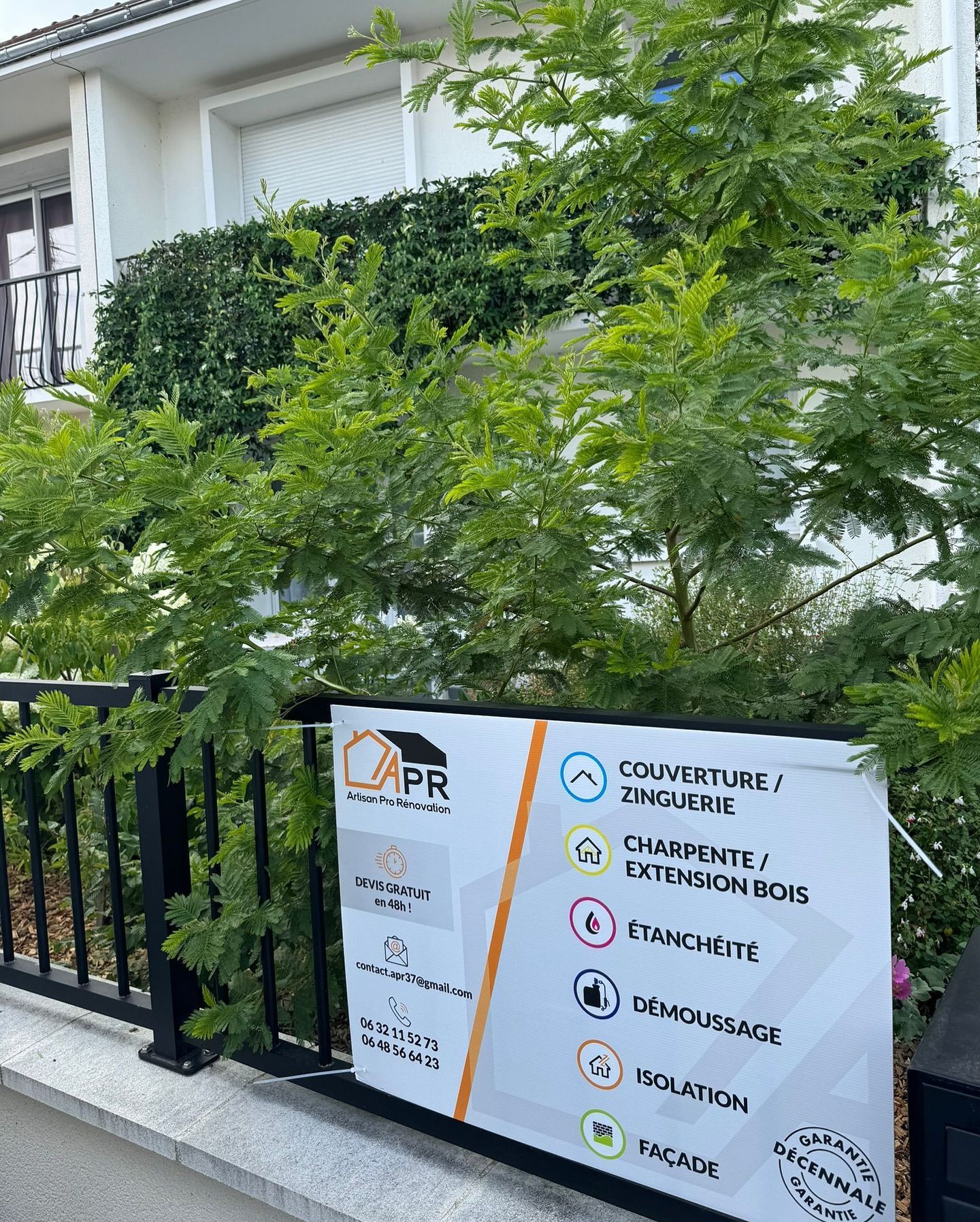 Panneau d'une entreprise de construction devant une maison avec des arbres et des buissons verts.