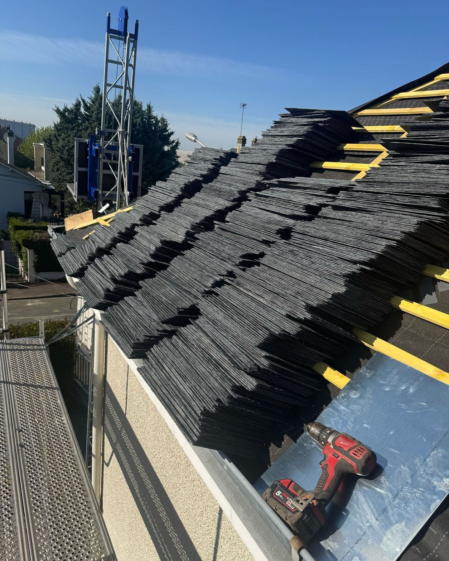 Toit avec des piles de bardeaux noirs, des outils de construction et une tour bleue en arrière-plan.