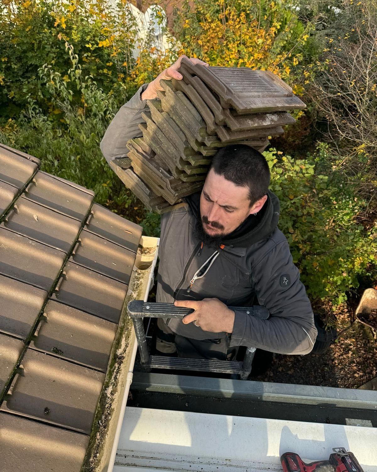 Un couvreur sort d'un accès au toit en portant des tuiles, regardant vers le bas. Des plantes vertes apparaissent en arrière-plan.