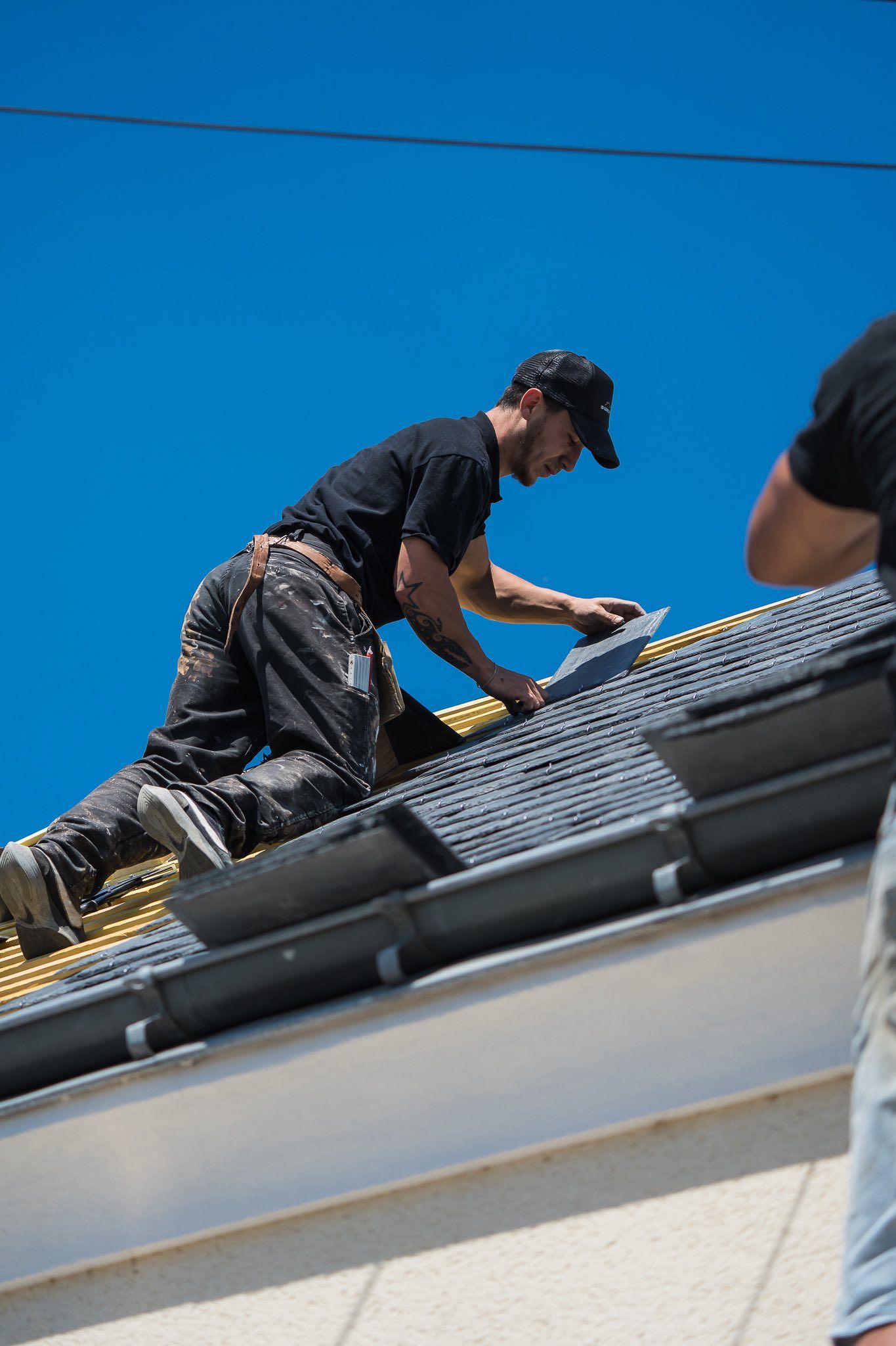 Couvreur en chemise et casquette noires, agenouillé sur un toit, installant des tuiles. Ciel bleu éclatant.
