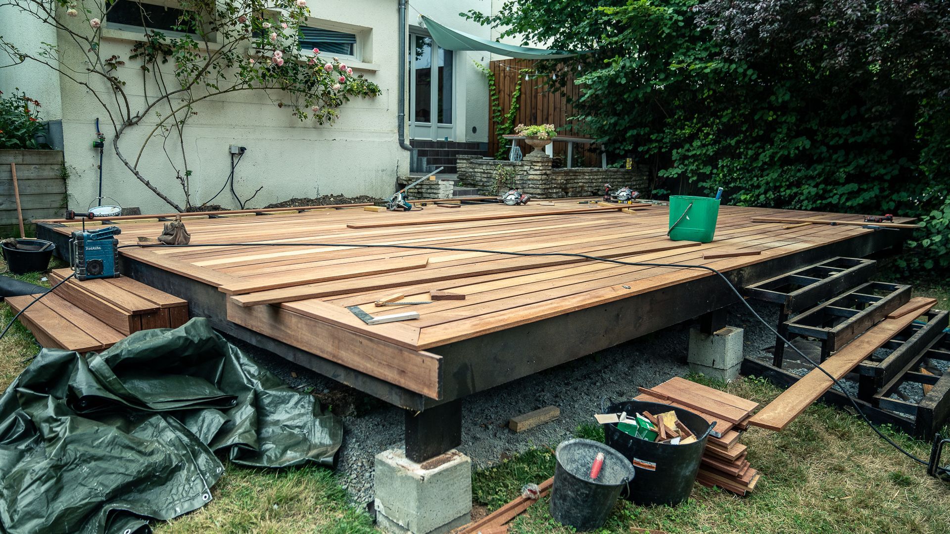 Construction d'une terrasse en bois dans un jardin. Partiellement terminée, avec outils et matériaux dispersés un peu partout.
