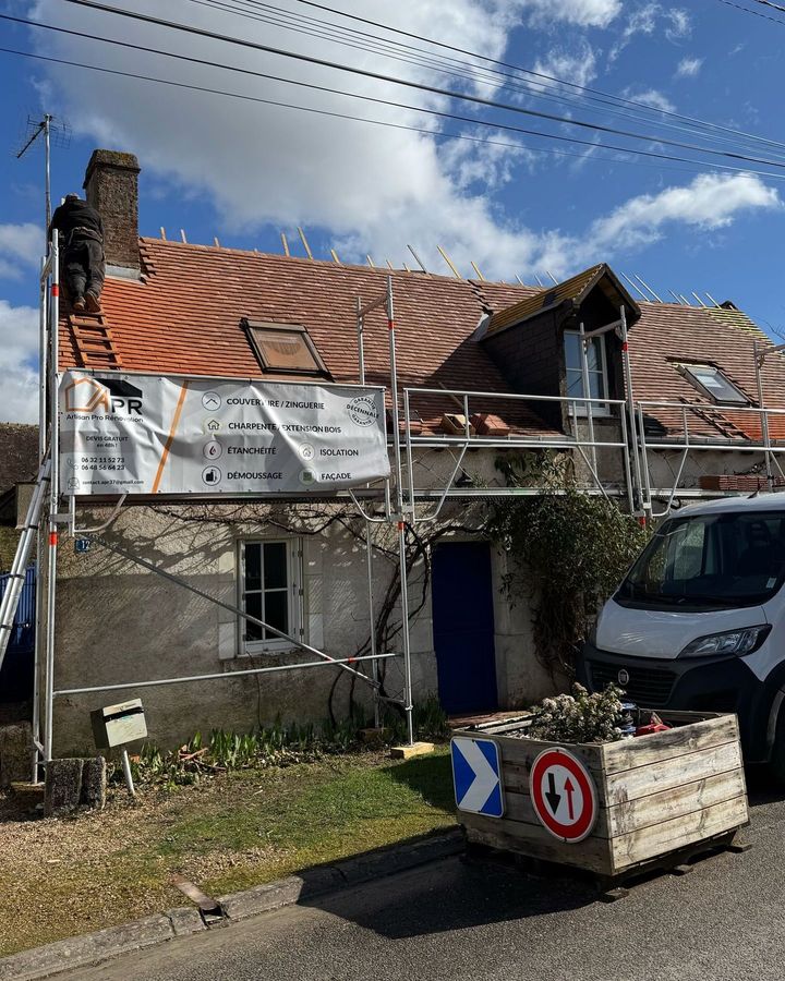 Maison en construction avec échafaudage, ouvrier sur le toit. Toiture en tuiles rouges, murs en pierre grise.