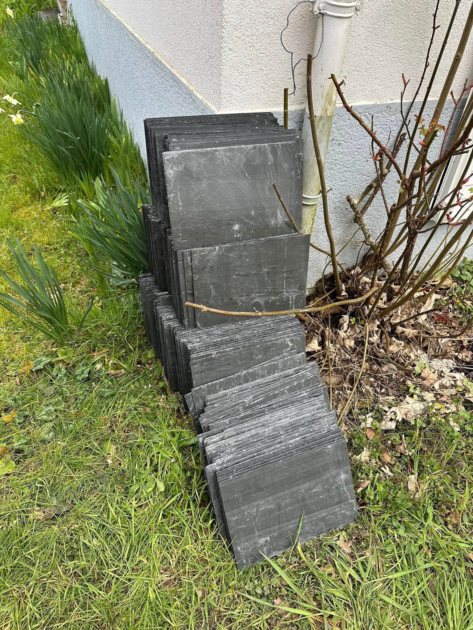 Une pile de tuiles gris foncé appuyées contre un mur et un tuyau fin, dans une zone herbeuse.