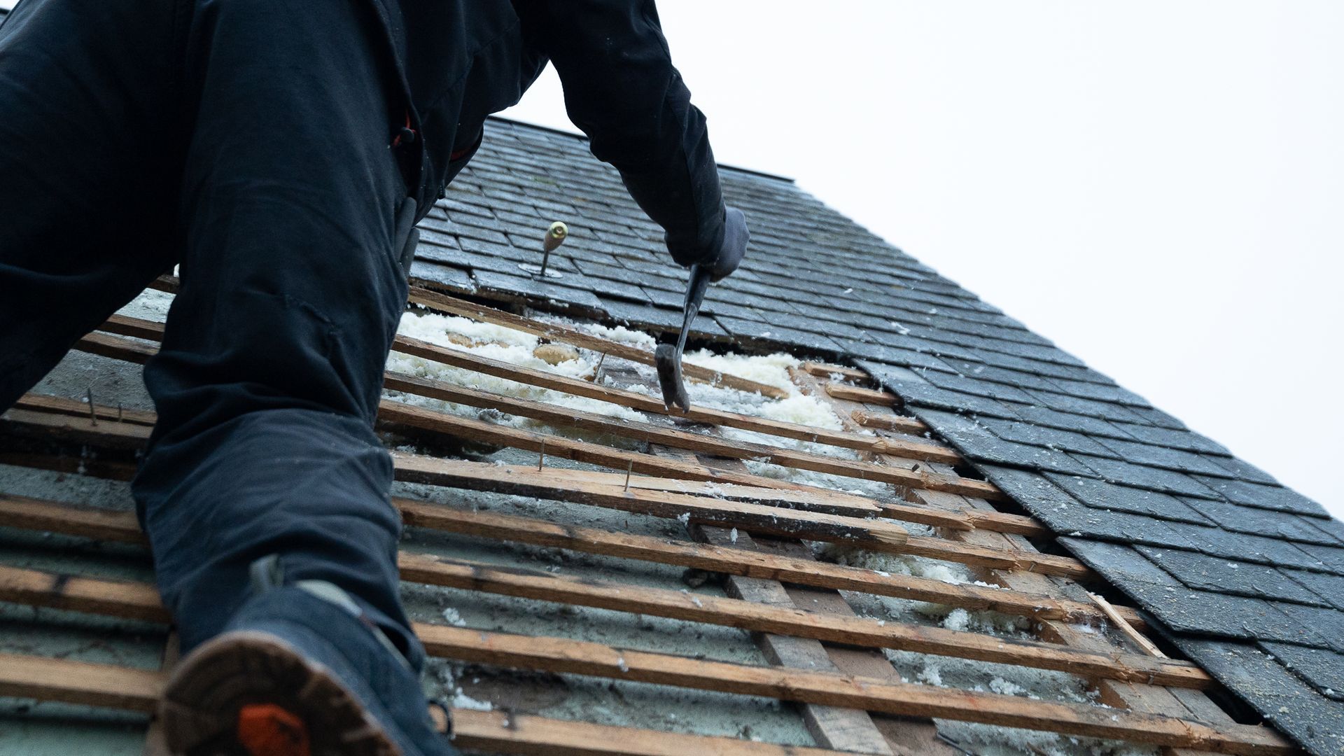 Personne sur un toit, enlevant des bardeaux avec un outil, révélant la structure en bois et l'isolation en dessous.