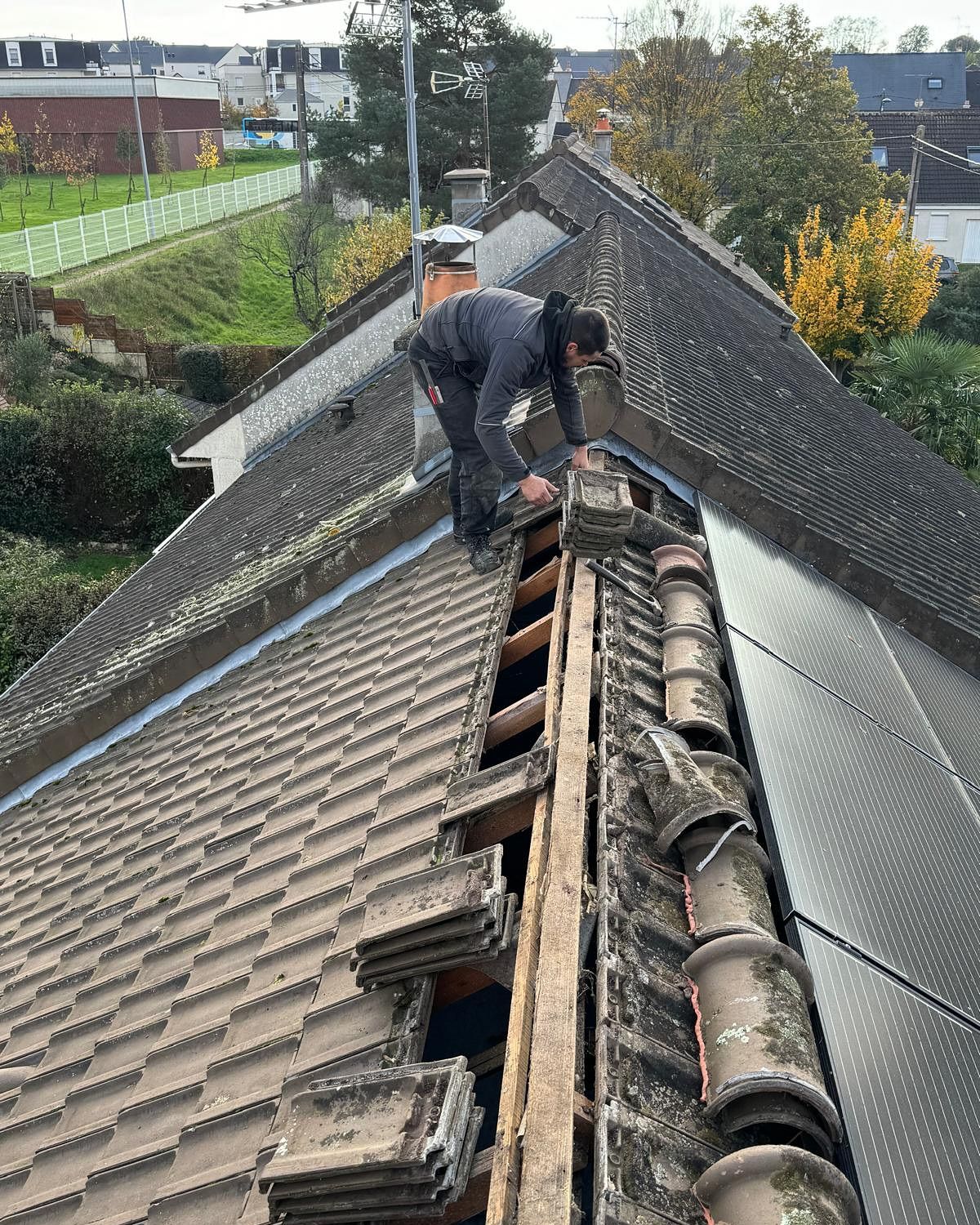 Couvreur enlevant des tuiles sur un toit en pente. Tenue grise, extérieur du bâtiment.