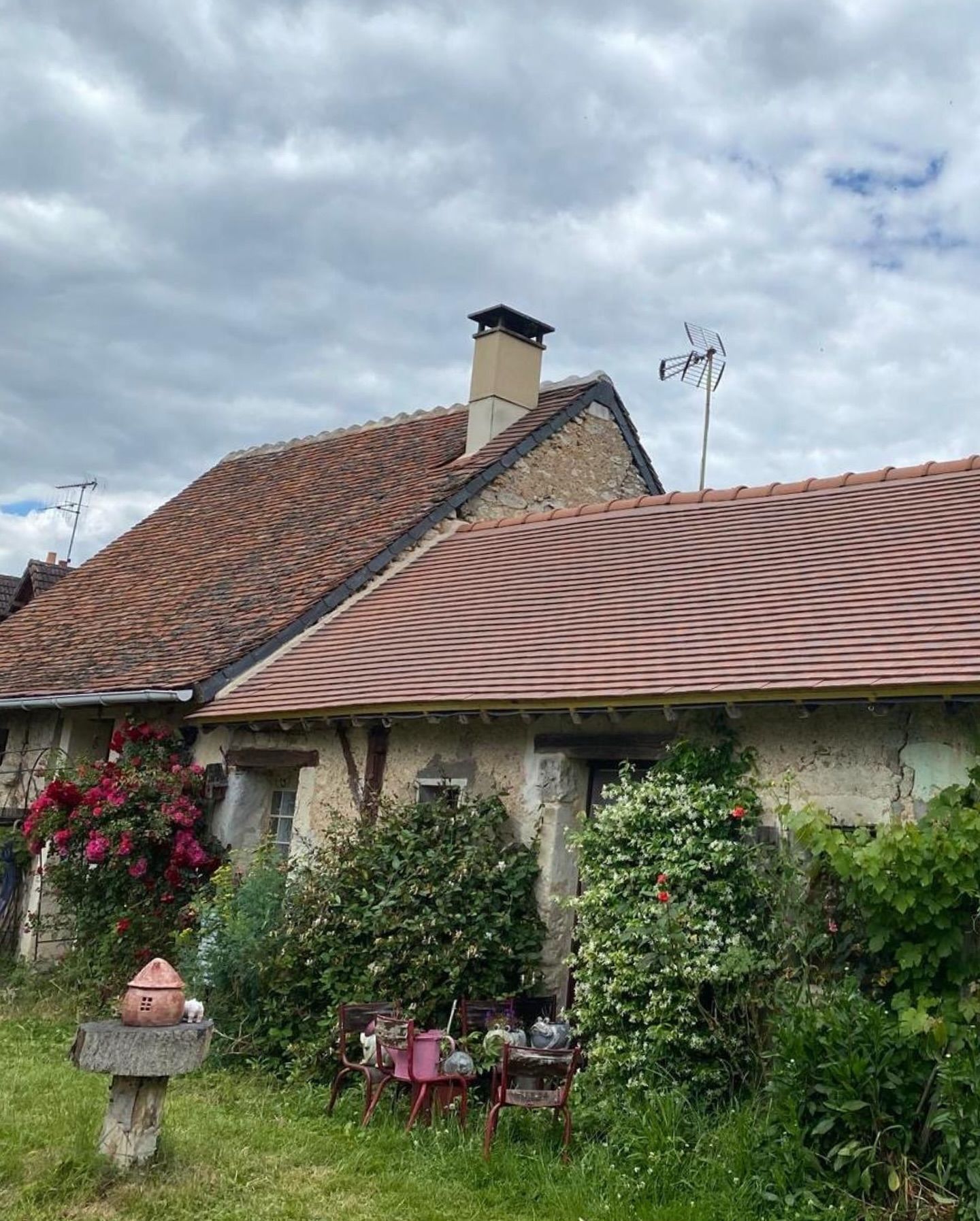 Maison en pierre aux toits de tuiles rouges, entourée de verdure et d'un ciel nuageux.