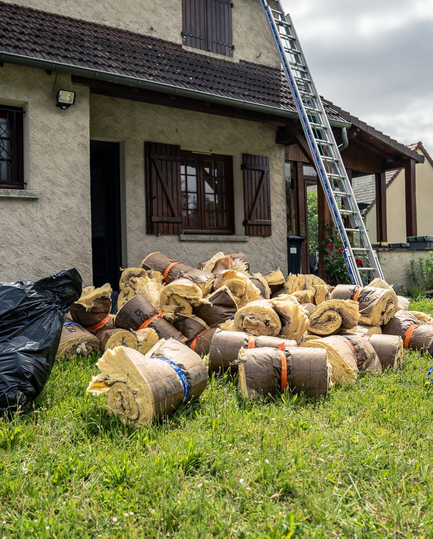 Rouleaux d'isolant sur l'herbe près d'une maison avec une échelle, suggérant une amélioration de la maison.