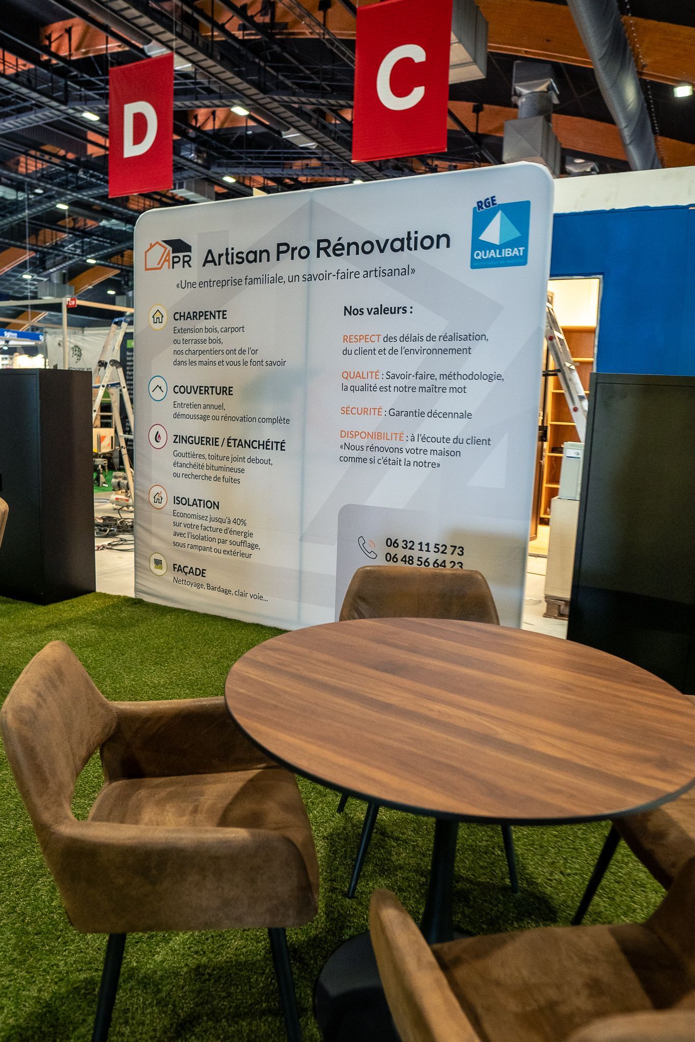 Un stand d'exposition avec une table et des chaises devant une affiche, un sol en gazon.