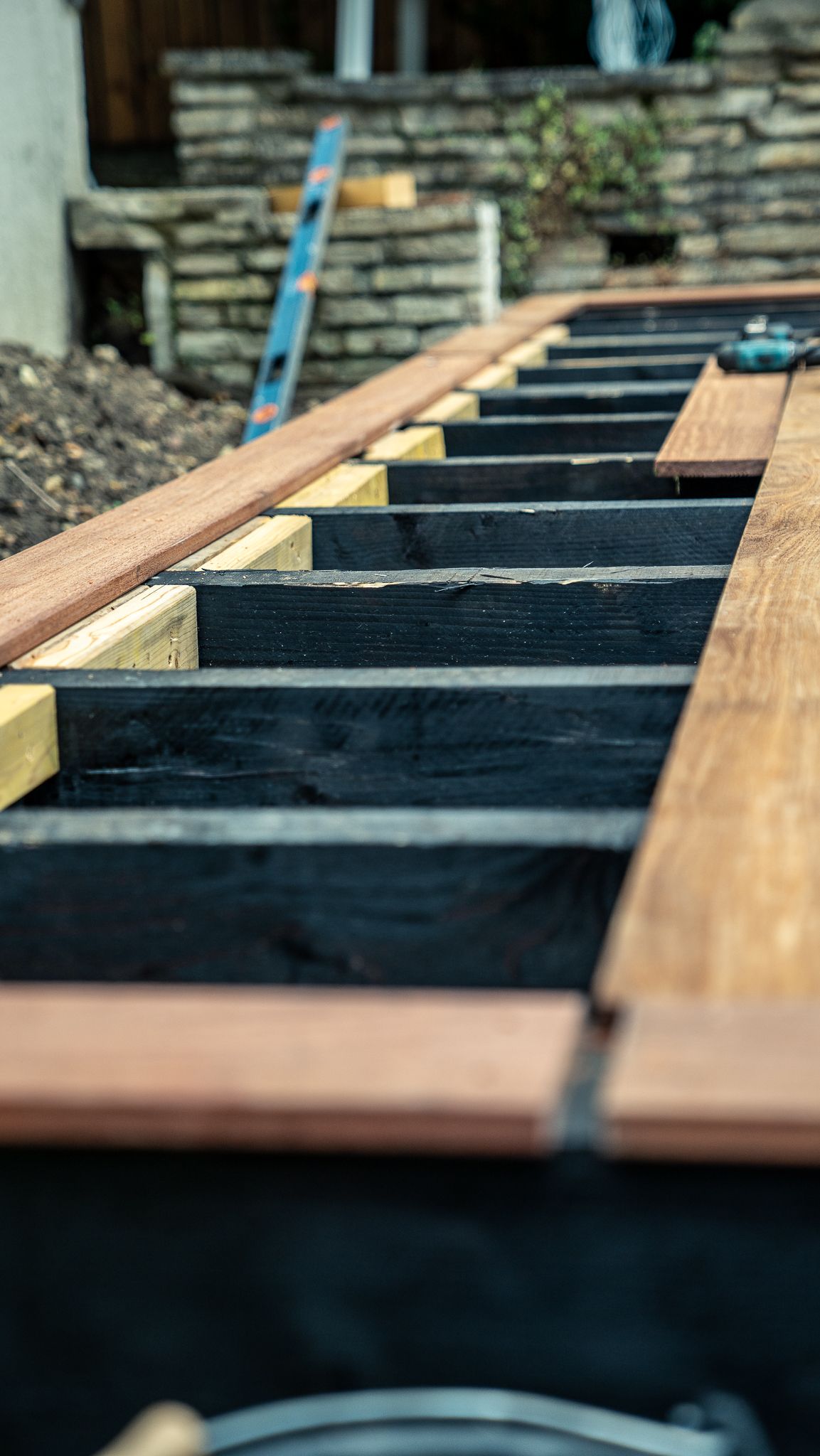 Terrasse en construction, poutres foncées supportant des planches marron. Construction dans une cour.