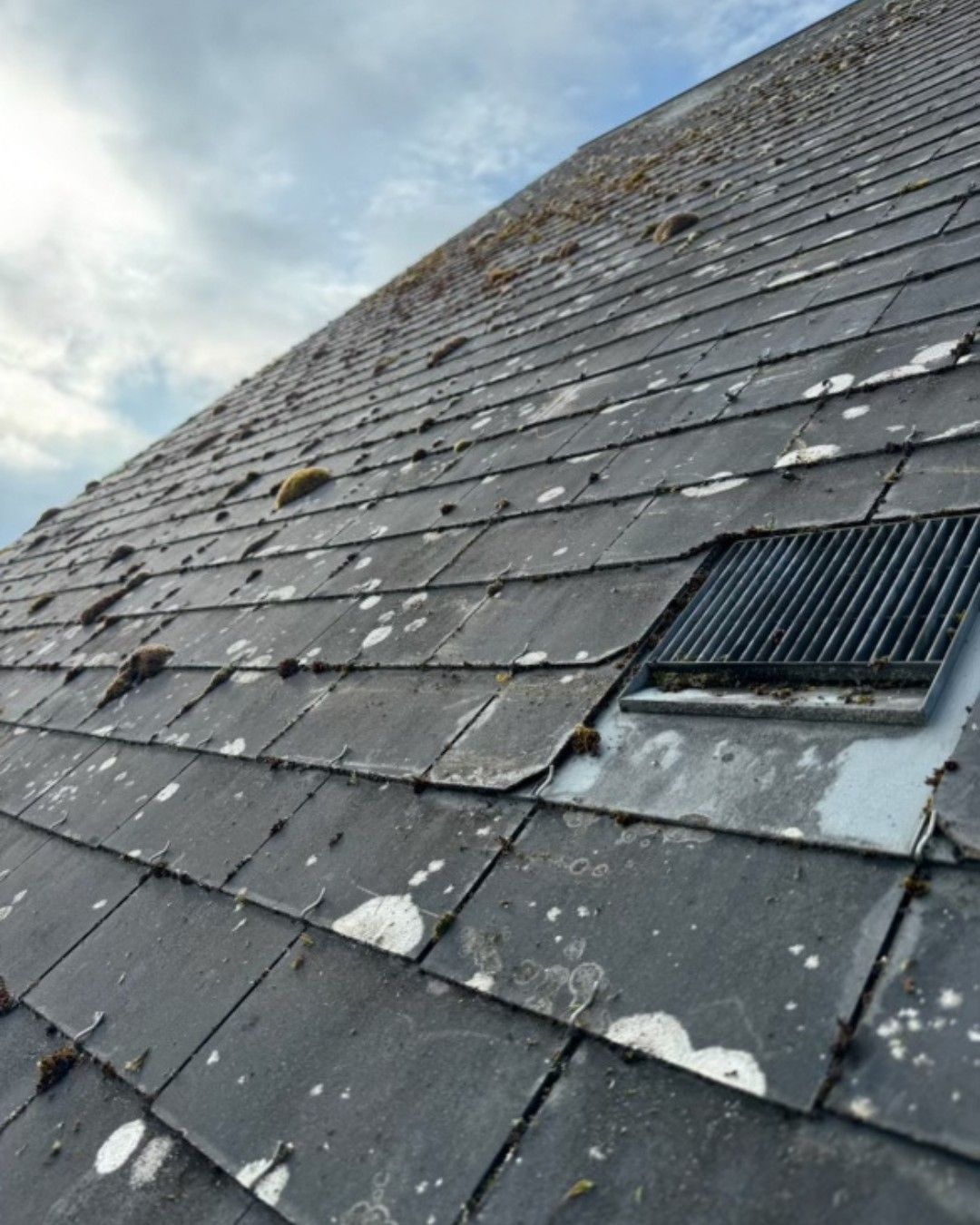 Vue en angle d'un toit en ardoise avec des plaques de mousse et une grille d'aération. Ciel nuageux.