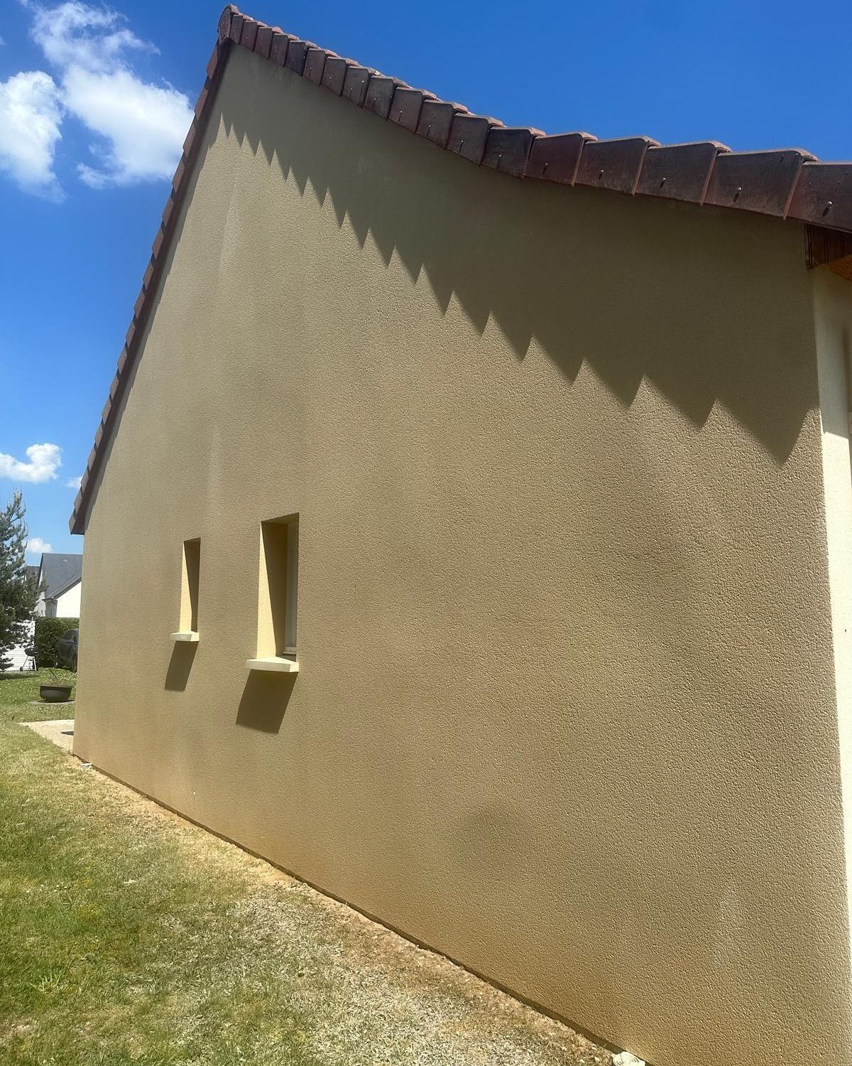 Mur en stuc beige avec deux fenêtres, toit en tuiles rouges et cour herbeuse sous un ciel bleu.