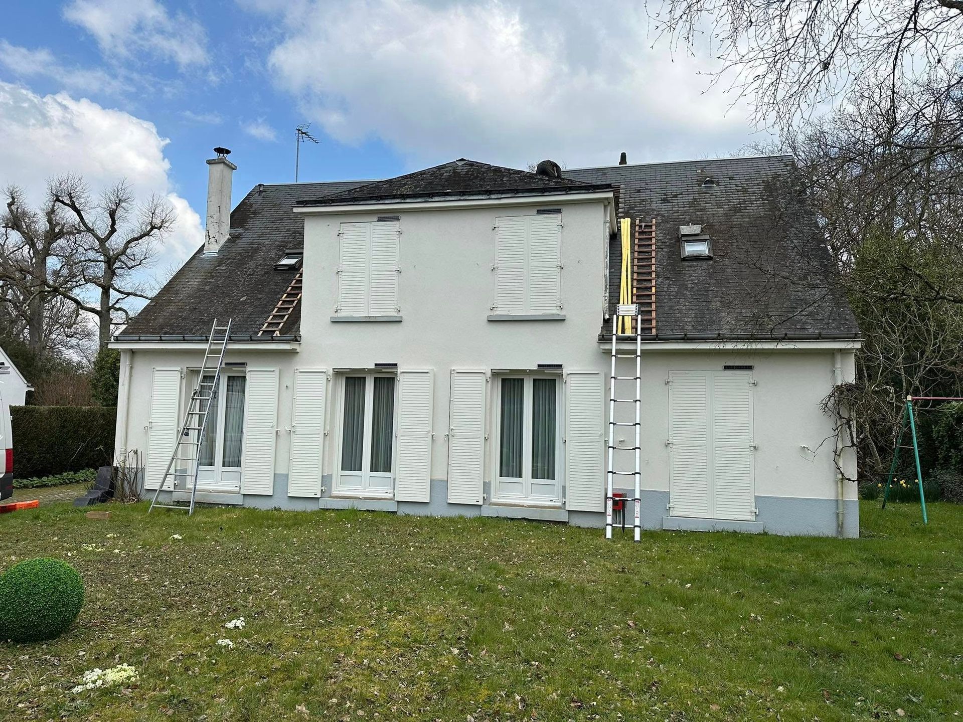 Extérieur de la maison avec revêtement blanc, volets et toiture partiellement remplacée, échelles présentes.