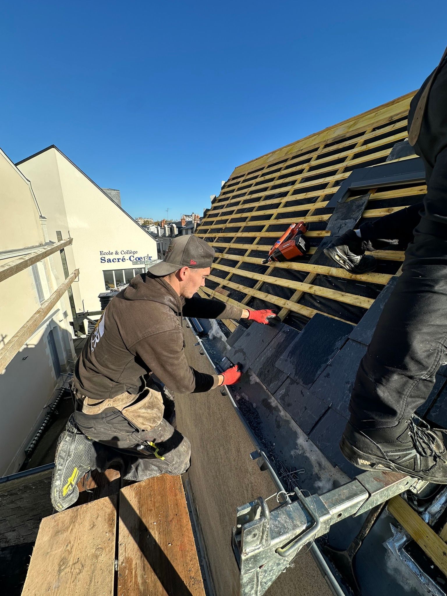 Couvreur installant des bardeaux sur un toit en pente sous un ciel bleu éclatant.