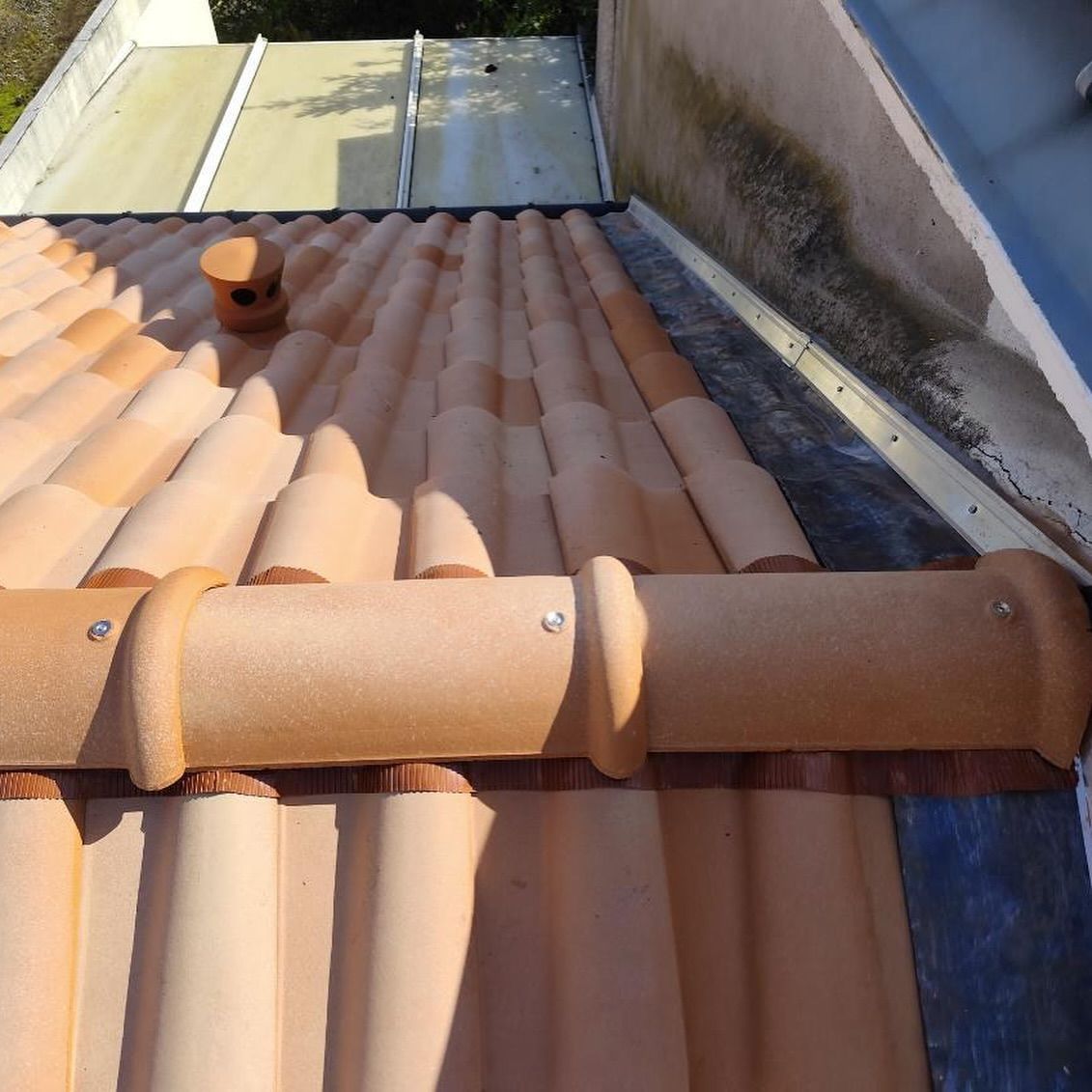 Toiture en tuiles d'argile avec gouttière, tuiles marron clair, petite cheminée et mur blanc adjacent.