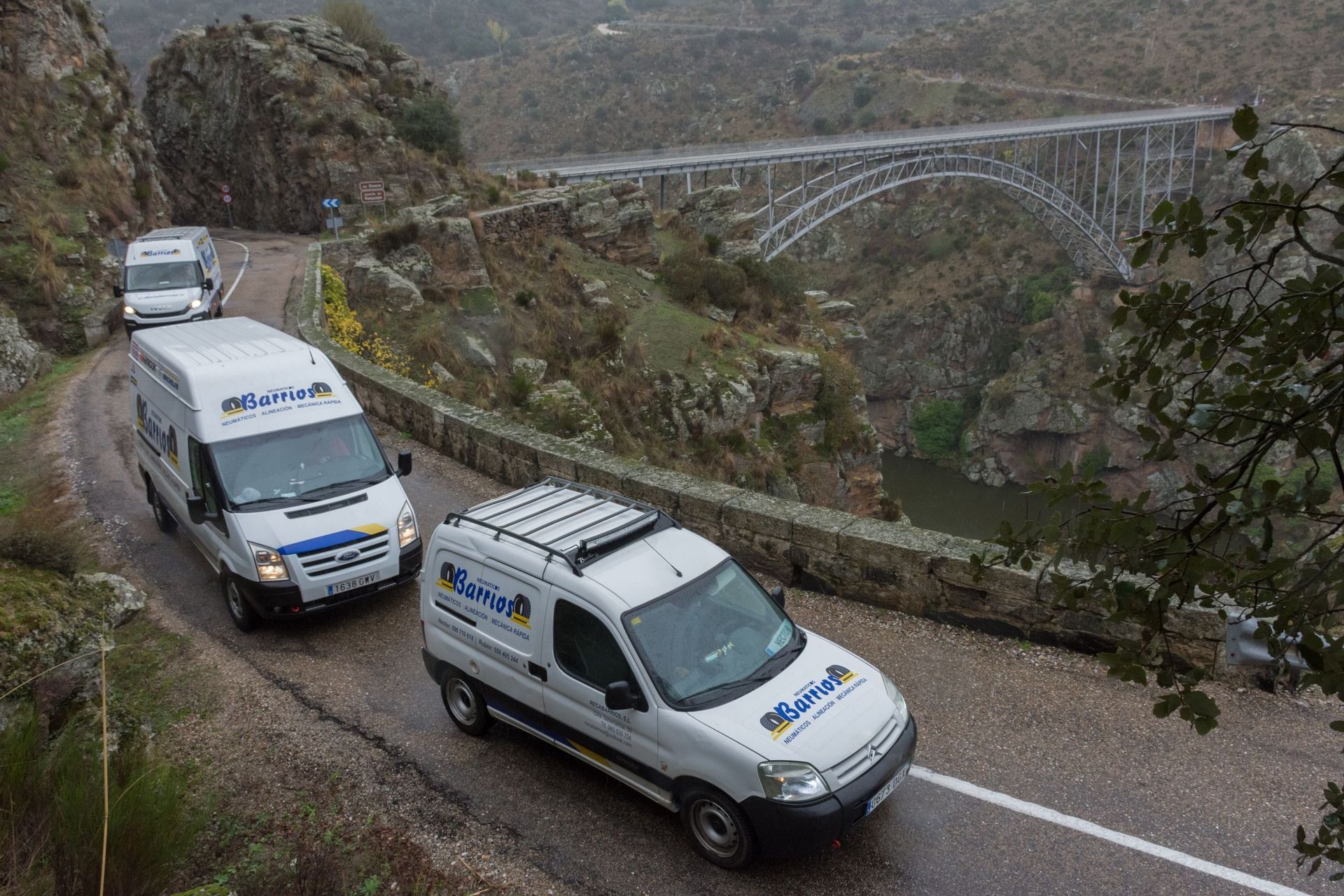 Tres furgonetas blancas en una carretera sinuosa con un puente al fondo, situada en una zona montañosa.
