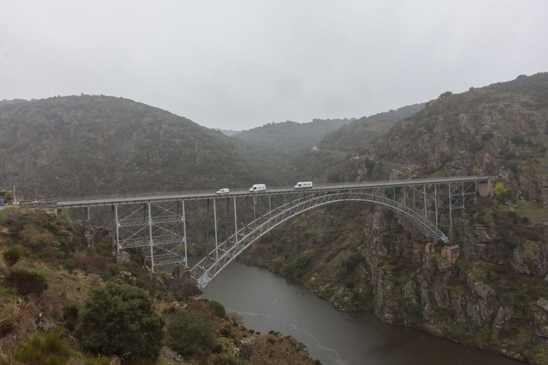 Puente de arco de acero sobre un río, con vehículos cruzando. Paisaje montañoso, cielo nublado.