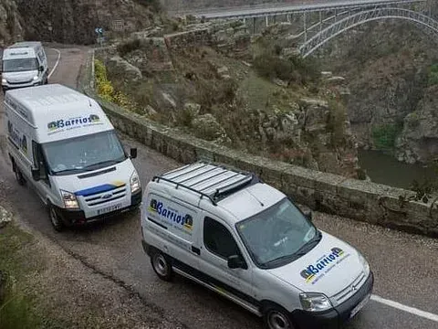 Tres furgonetas blancas circulando por una carretera de montaña con un puente al fondo. Llevan un logotipo.
