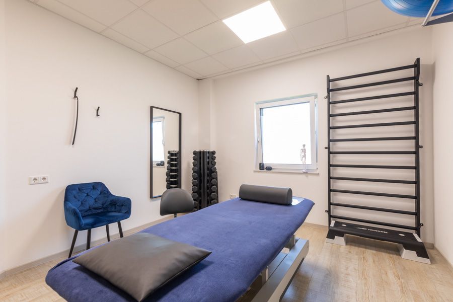 Heller Behandlungsraum mit Massageliege, blauem Stuhl, Wandspiegel und Übungsstangen.