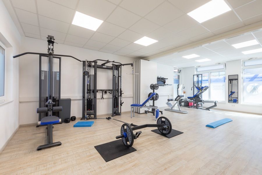 Heller Fitnessraum mit Trainingsgeräten, Gewichten und Matten auf hellem Holzboden.