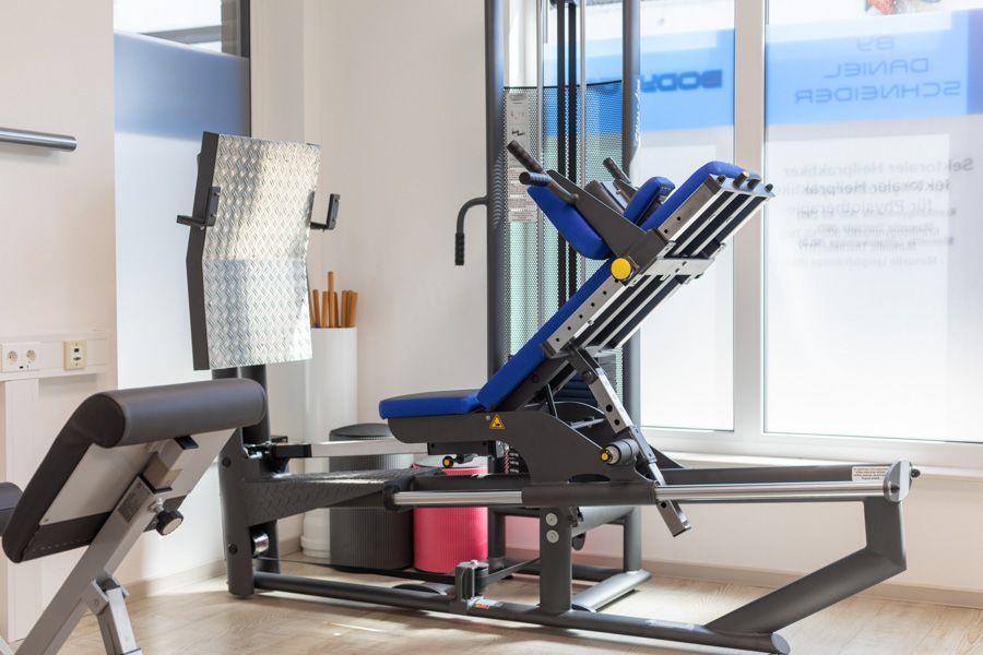Reformer-Pilates-Gerät in einem hellen Studio in der Nähe großer Fenster, schwarz-blauer Rahmen mit Gurten und Griffen.