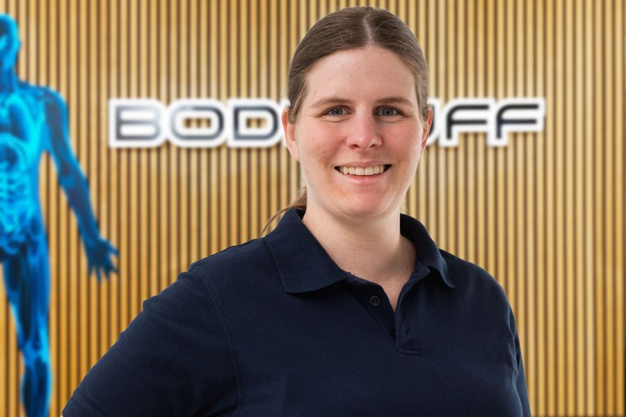 Eine lächelnde Person in einem dunkelblauen Polohemd steht vor einer Holzwand, auf der ein teilweise sichtbares Schild mit der Aufschrift „BODY…“ und die blaue Silhouette einer Figur zu sehen sind.
