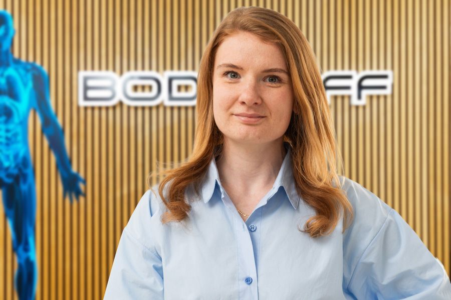 Eine Frau in einem hellblauen Hemd steht vor einer gestreiften Wand; hinter ihr ist ein Logo zu sehen.