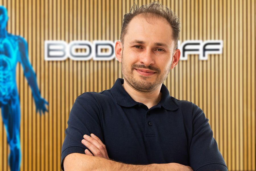 Ein Mann steht mit verschränkten Armen vor einer holzgetäfelten Wand mit Firmenlogo und trägt ein dunkelblaues Polohemd.