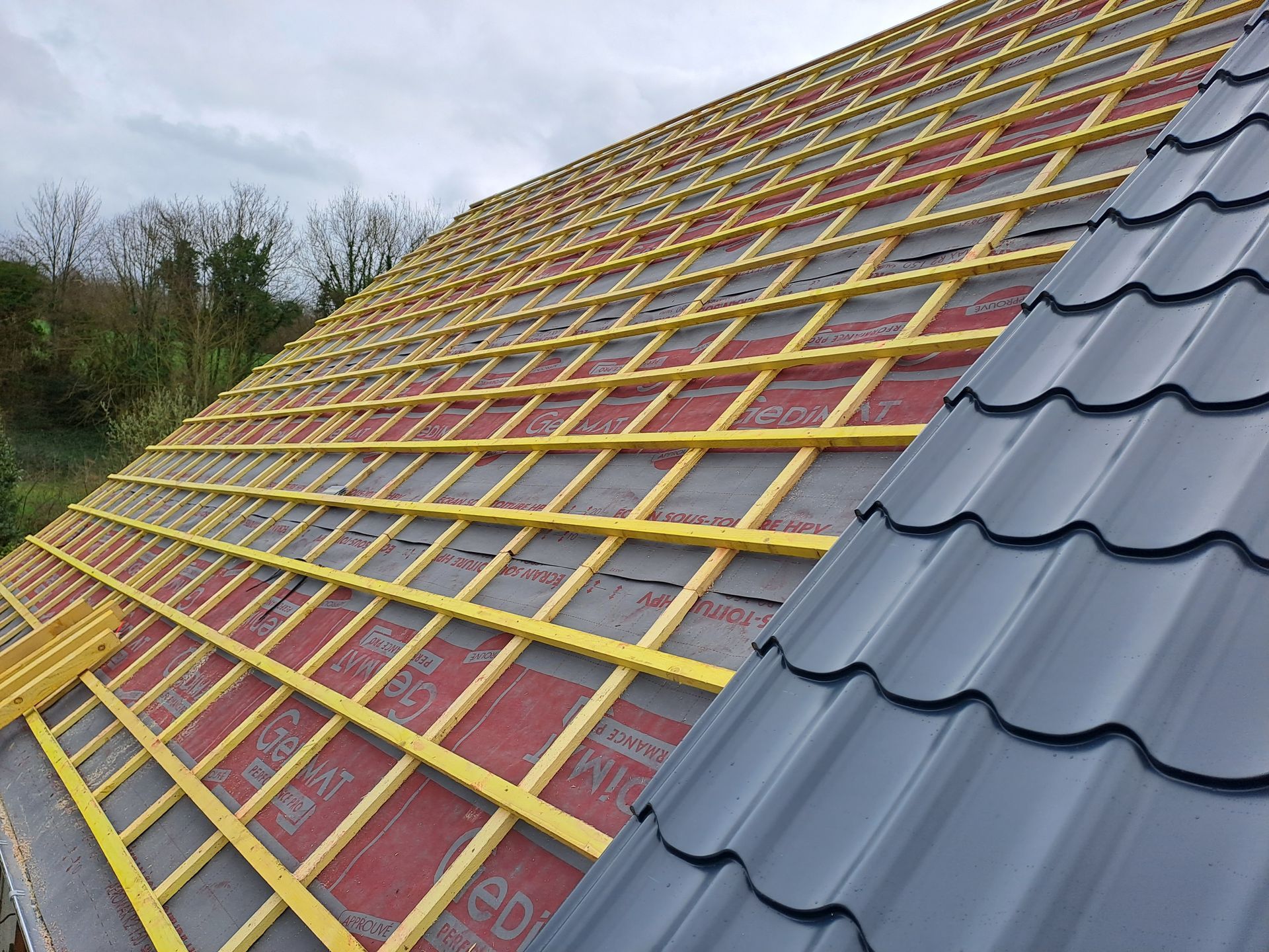 Toiture en construction avec charpente en bois, membrane rouge et tuiles métalliques grises.