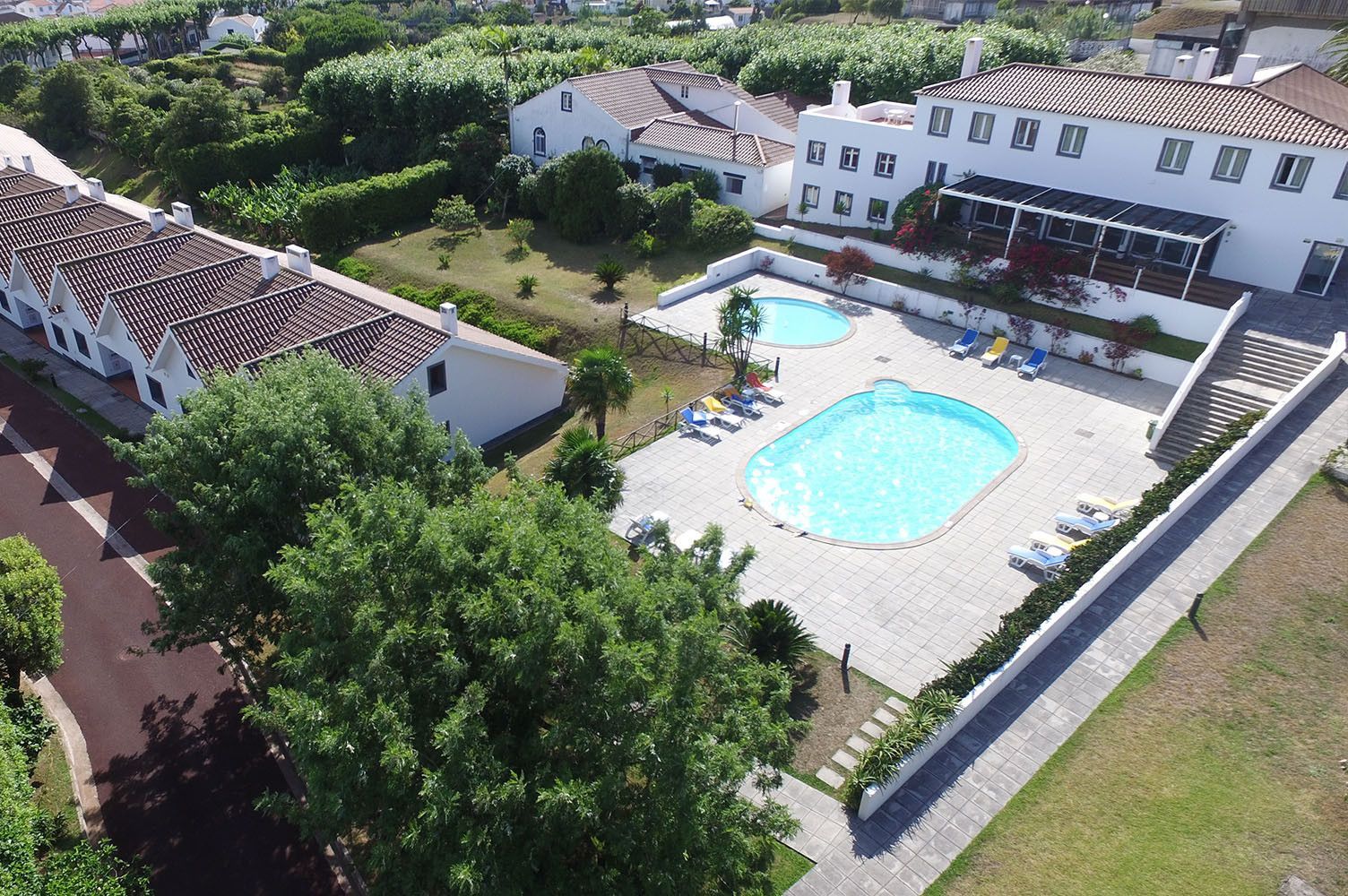 Vista aérea de um hotel branco com telhados de telha vermelha, duas piscinas e vegetação ao redor.