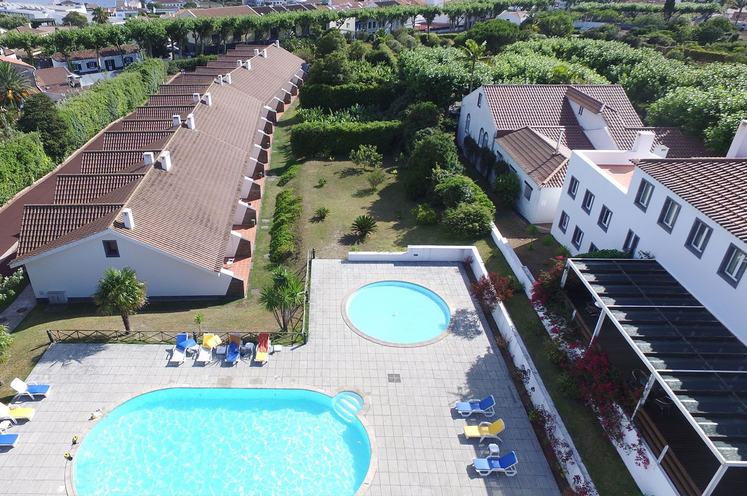 Vista aérea de um resort com duas piscinas, edifícios brancos e vegetação ao redor.