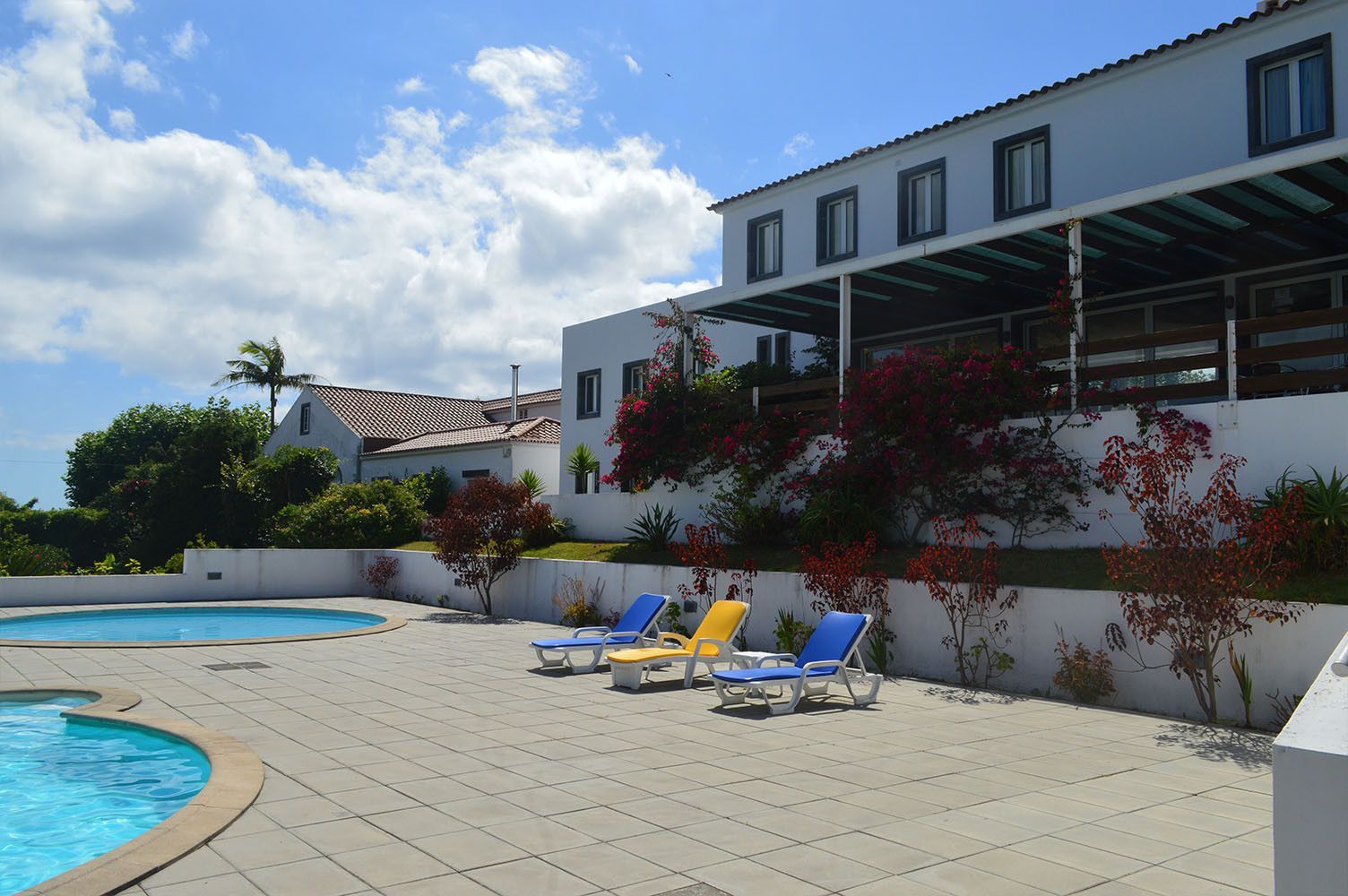 Piscina com espreguiçadeiras em frente a um prédio branco com arbustos floridos vermelhos sob um céu azul.