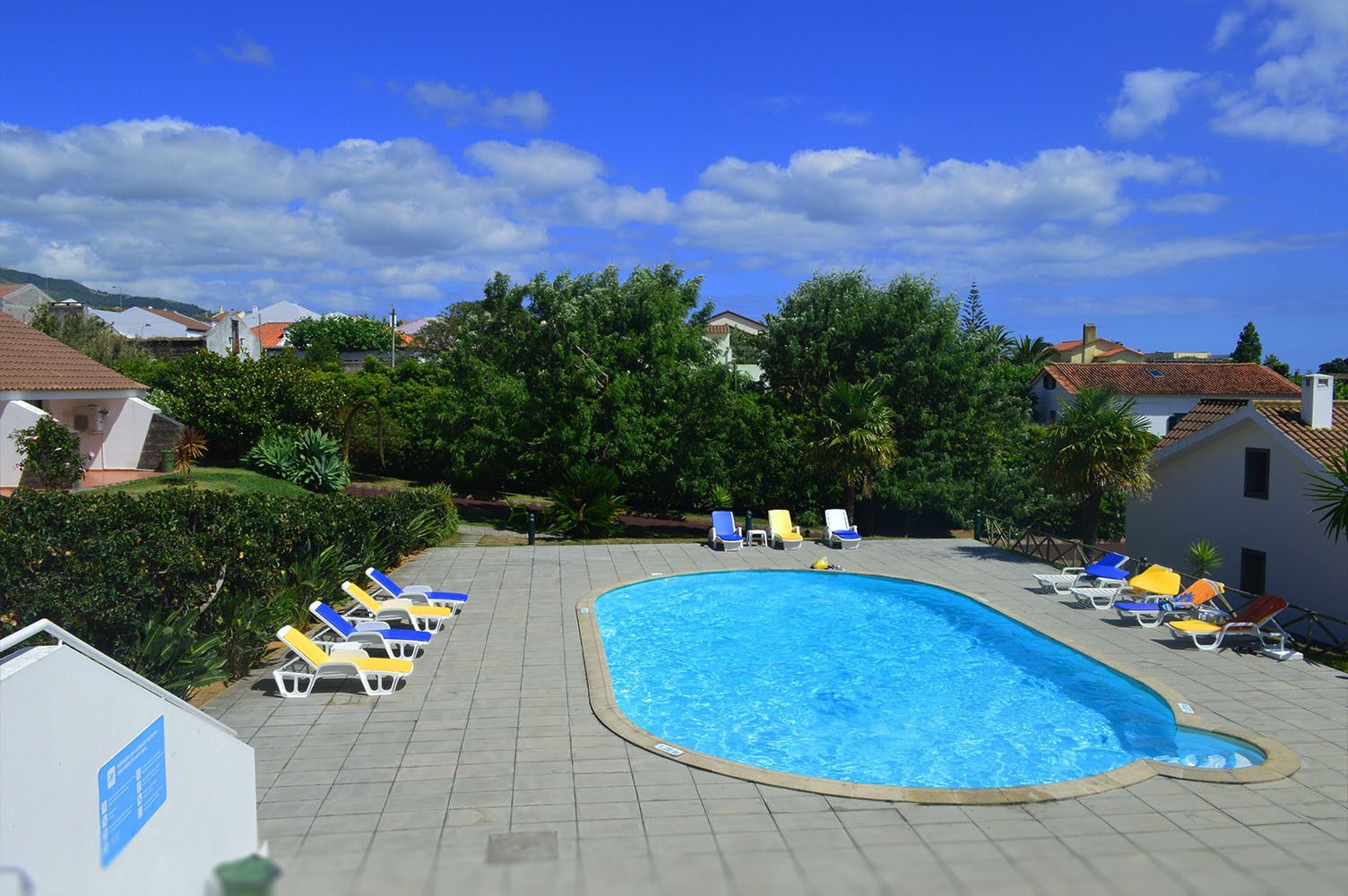 Piscina rodeada por espreguiçadeiras, árvores e edifícios, sob um céu azul.