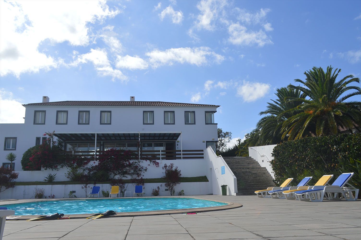 Prédio branco com piscina e espreguiçadeiras sob um céu azul com nuvens e palmeiras.