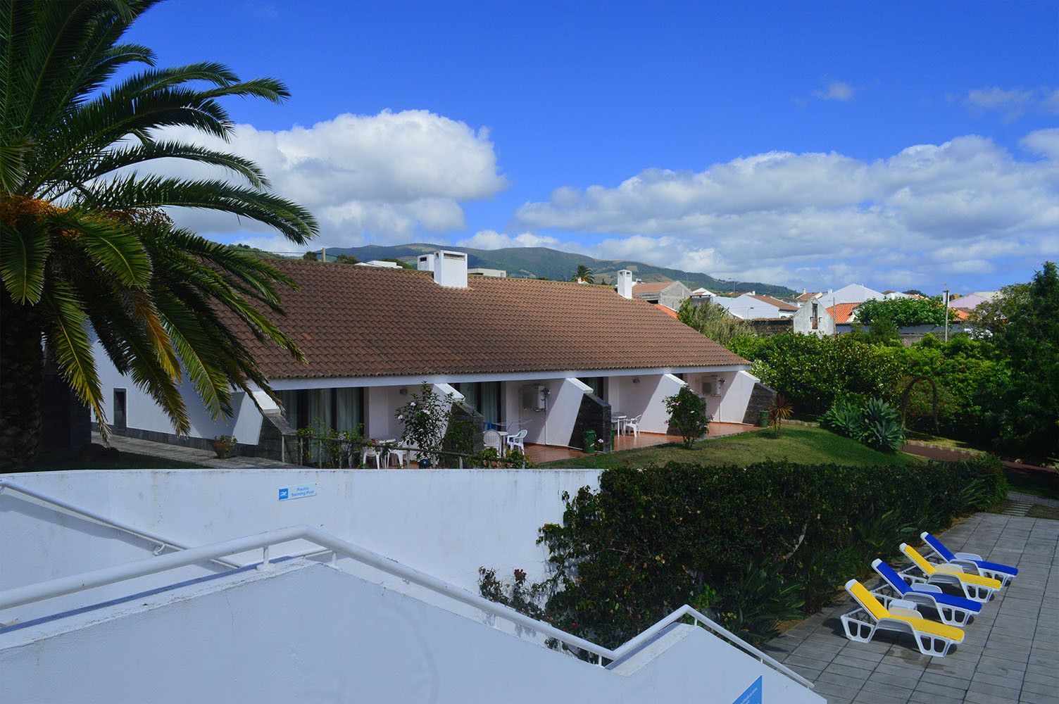 Prédios brancos com telhados marrons, palmeira, céu azul e espreguiçadeiras.