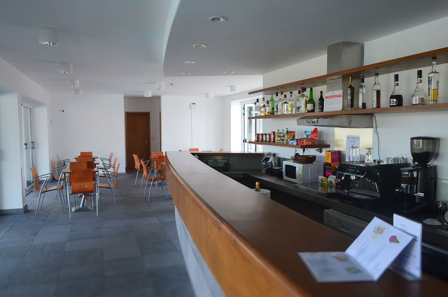 Área de bar com balcão de madeira, prateleiras de bebidas, mesas e cadeiras sobre piso de azulejos cinza.