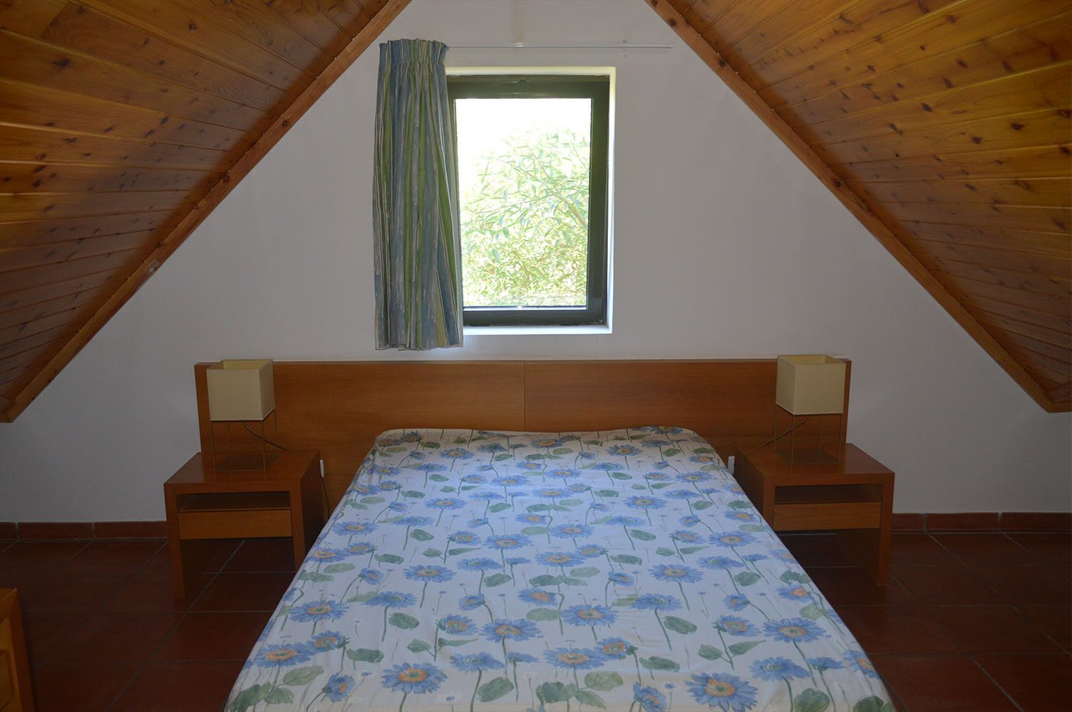 Quarto com cama, mesas de cabeceira e janela com cortina. Teto e cabeceira de madeira.