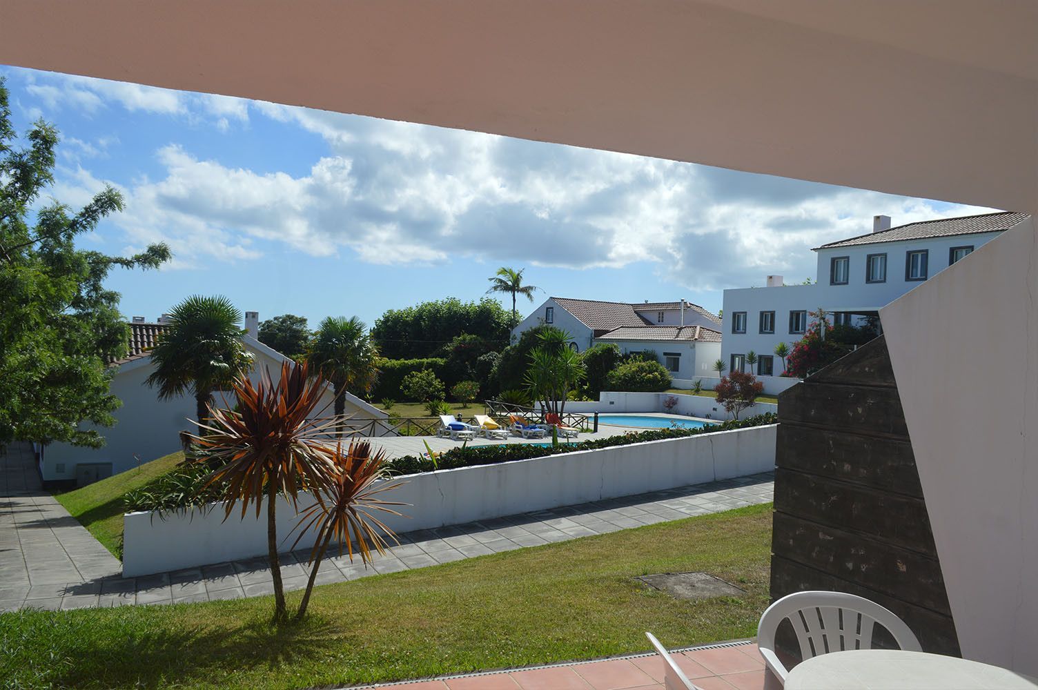 Vista da varanda de uma área de piscina com prédios, grama e árvores sob um céu azul com nuvens.