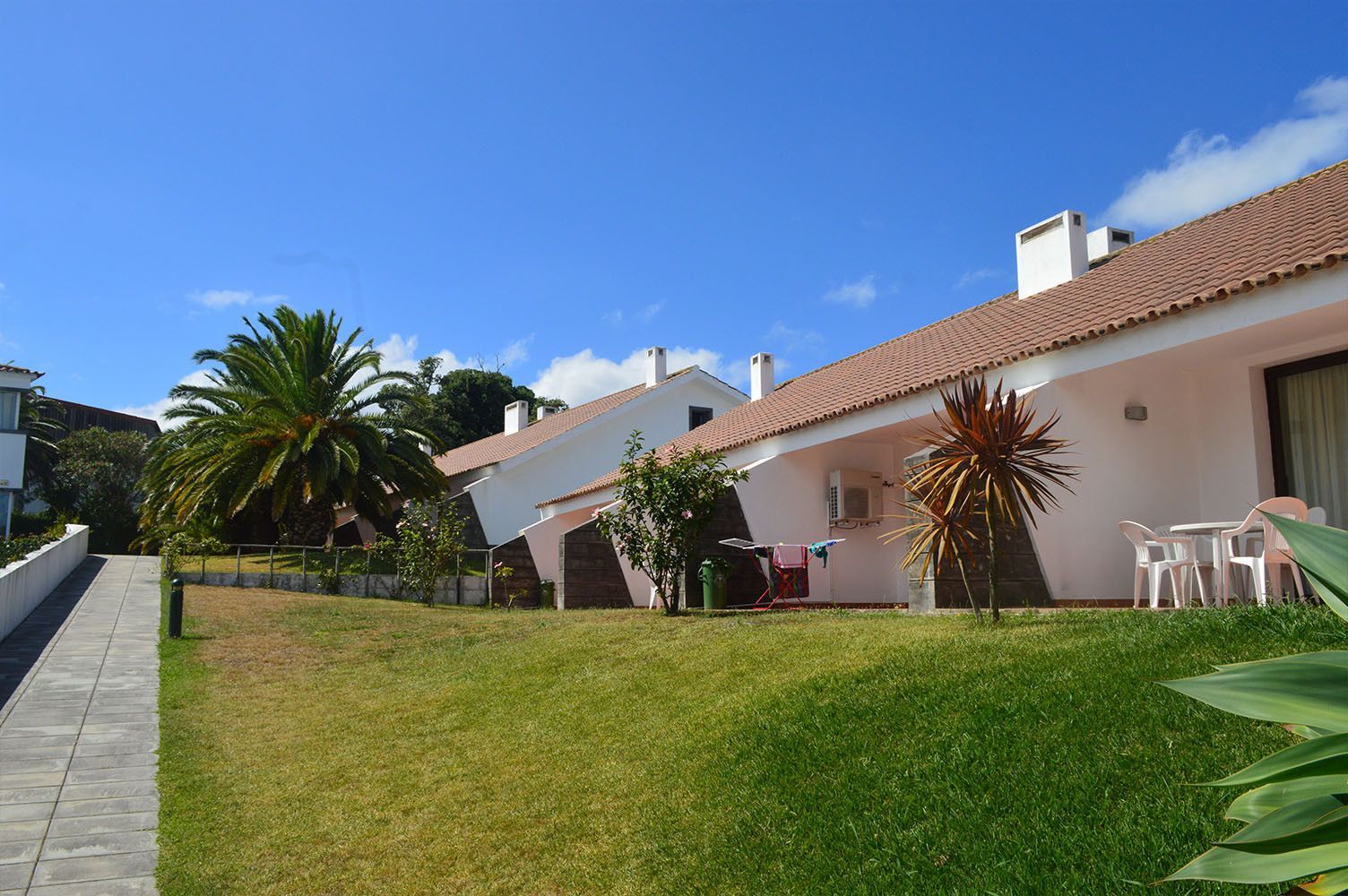 Fileira de prédios brancos com telhados de telha vermelha, grama verde e palmeiras sob um céu azul.