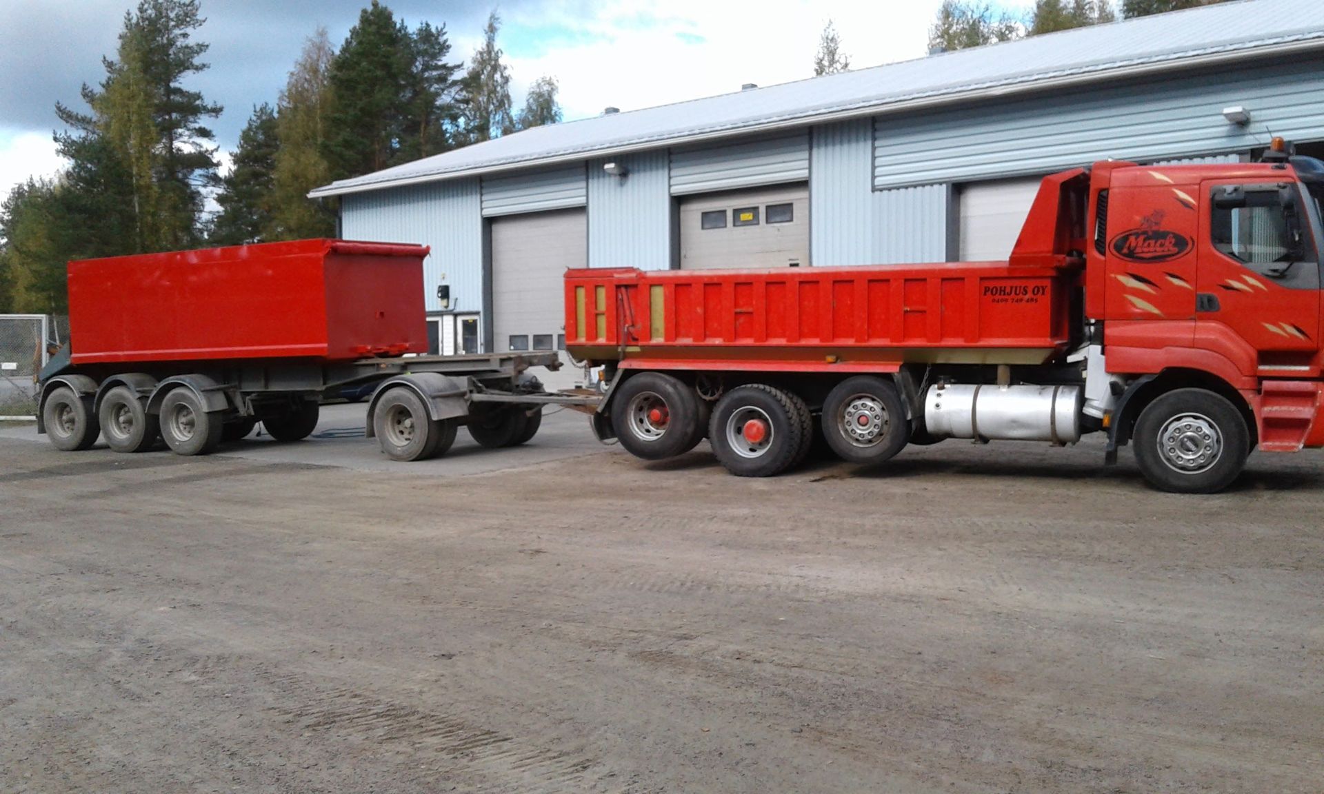 Punainen kippiauto ja perävaunu soratiellä, pysäköitynä rakennuksen eteen.