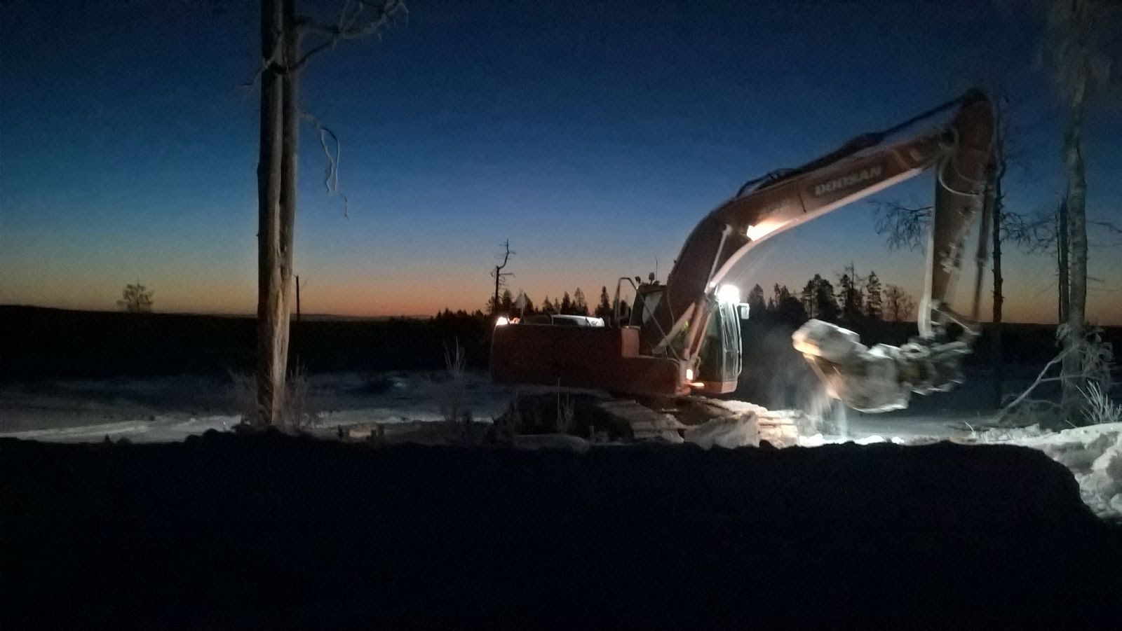 Yöllä työskentelevä kaivinkone, valojen valaistuna, koiran ollessa lähellä lumisella pellolla sinisen taivaan alla.
