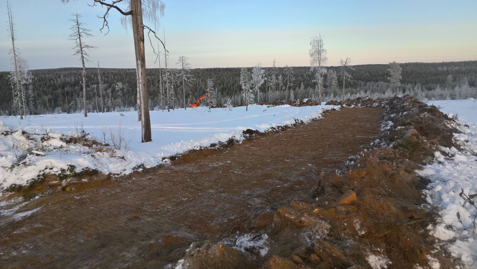 Luminen maisema, jonka etualalla on maakasa. Kaivinkone työskentelee etäällä, taustalla metsää.