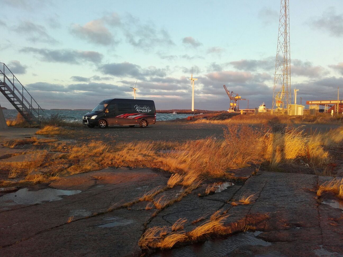 Musta pakettiauto pysäköitynä kallioiselle rannalle, jossa on teollisuusrakennuksia ja pilvinen taivas auringonlaskun aikaan.