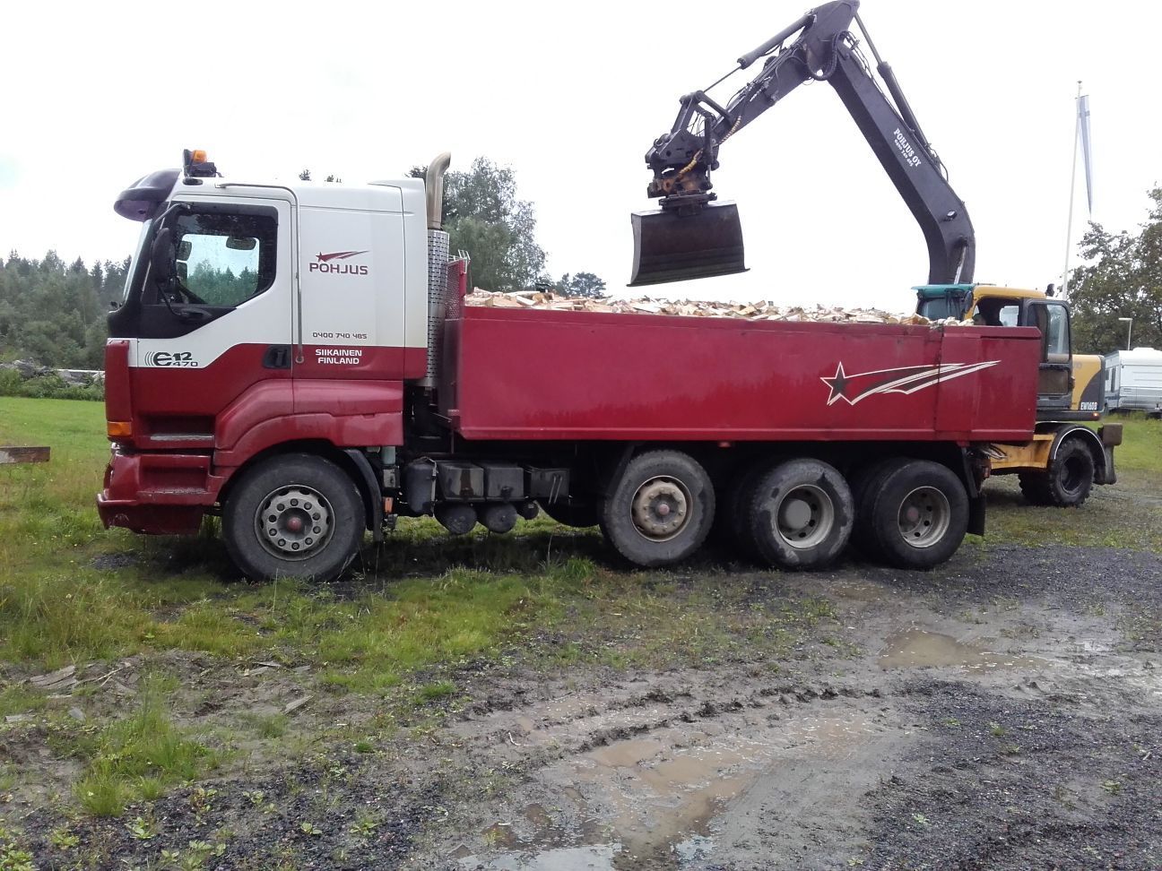 Punavalkoista kuorma-autoa lastataan materiaalilla kaivinkoneella ulkona mutaisella pellolla.