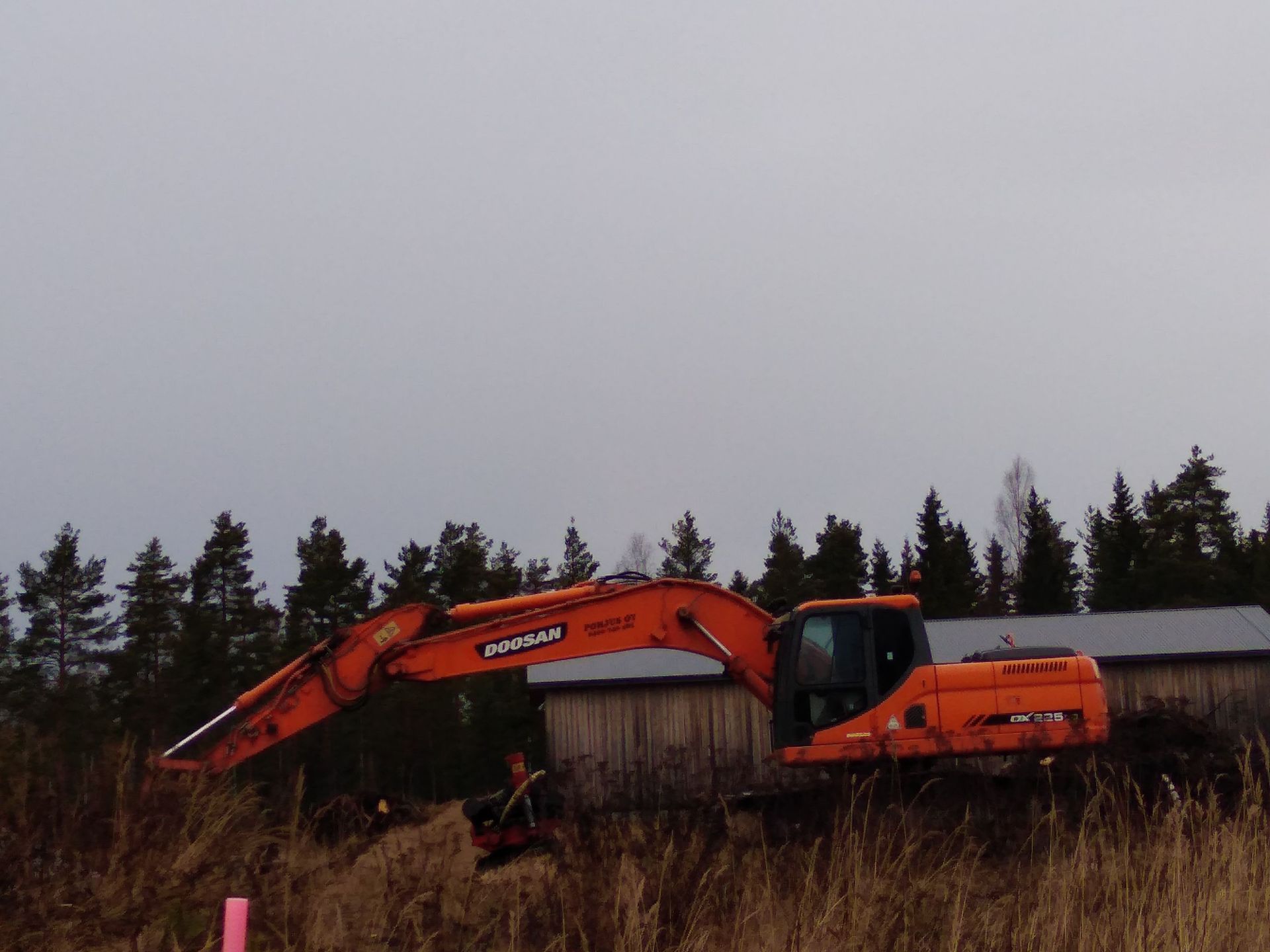 Oranssi kaivinkone työskentelee ulkona puiden ja rakennuksen lähellä pilvisenä päivänä.