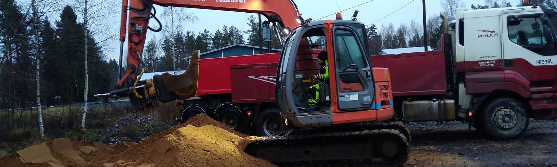 Kaivinkone kauhaa maata kuorma-auton vierestä metsäympäristössä.